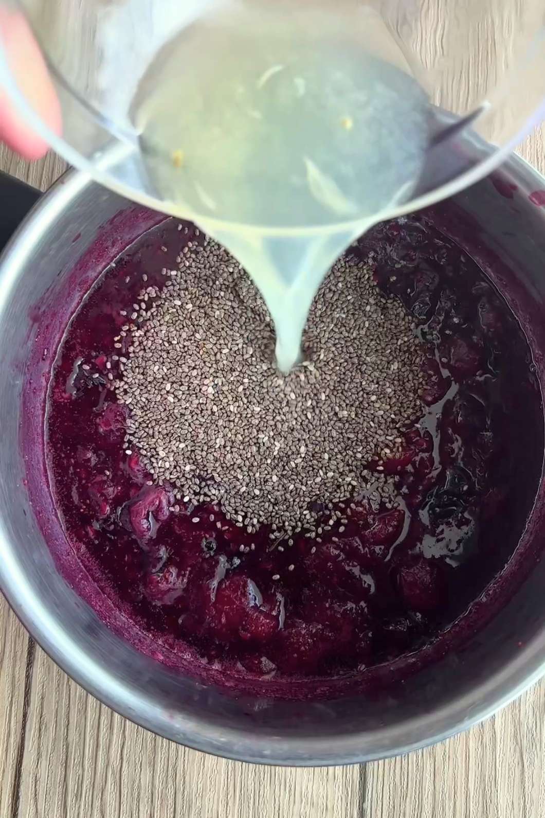 Hand pouring pale liquid over chia seeds atop dark raspberry jam in a metal bowl.