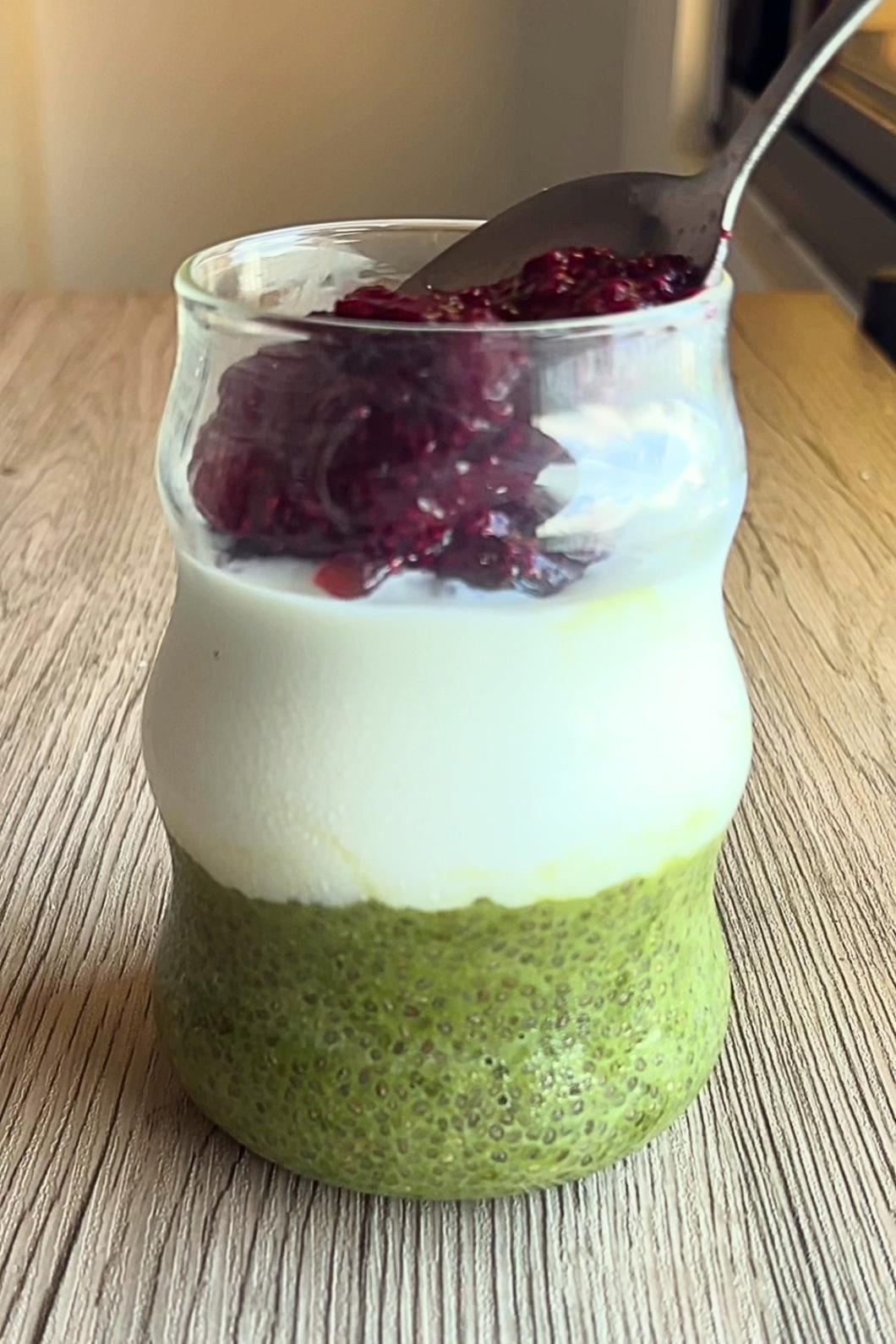 Spoon adding red jam atop white yogurt and green pudding layer in glass.