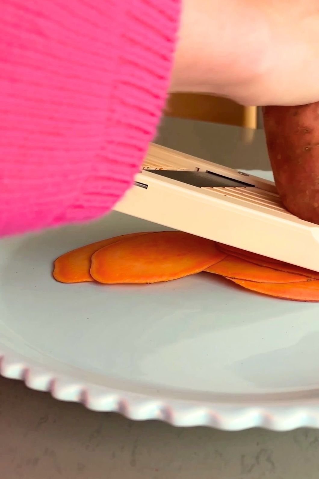 Hand slicing sweet potato on a mandolin, thin slices fanning out on a plate.