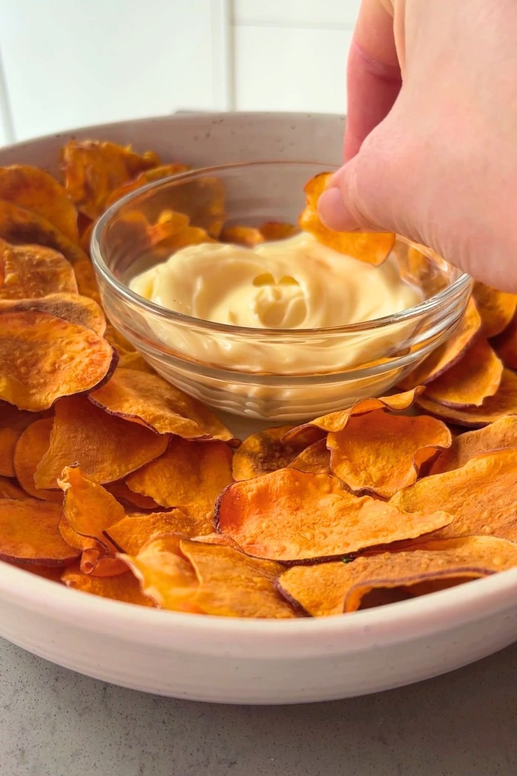 Hand dipping a crispy sweet potato chip into creamy dip in a clear glass bowl.