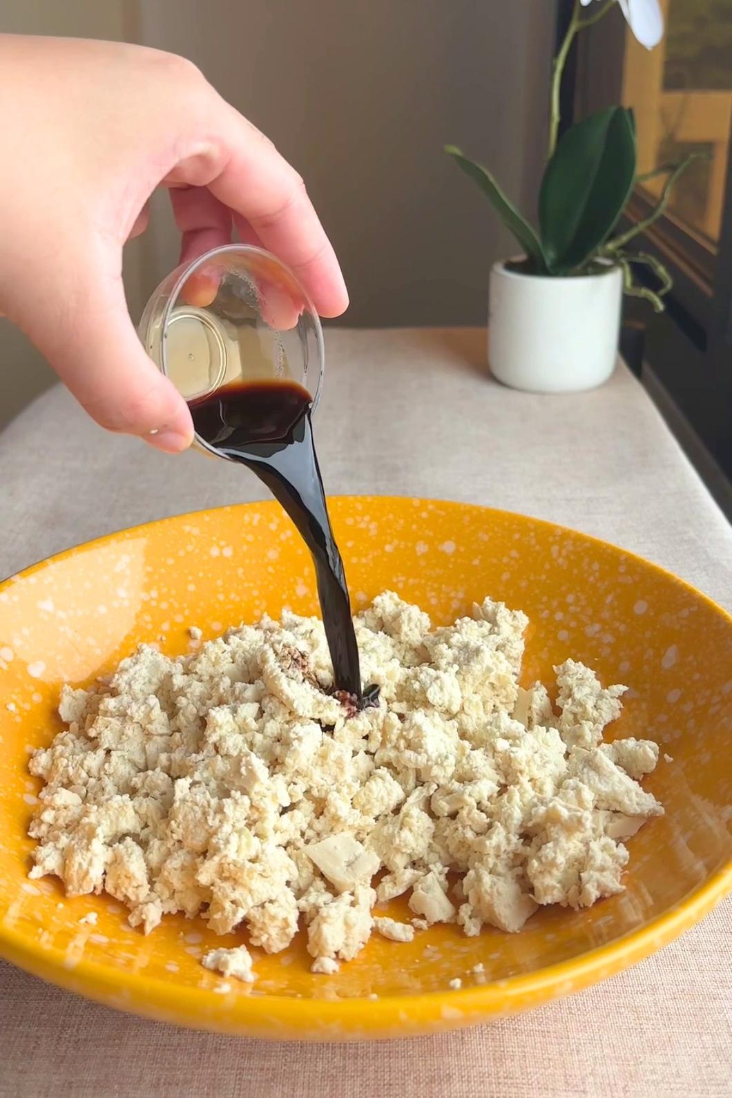 Hand pouring dark liquid from a small glass onto crumbled tofu in a bowl.
