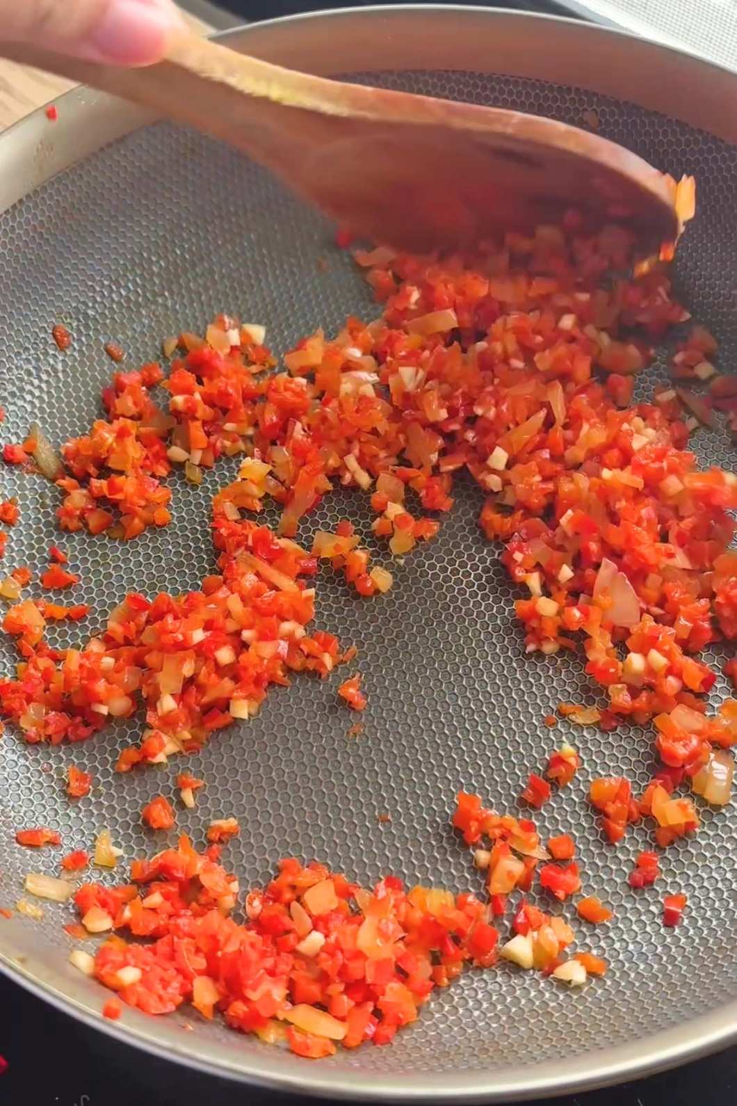 Wooden spoon stirring finely diced red capsicum and onion in a frying pan.