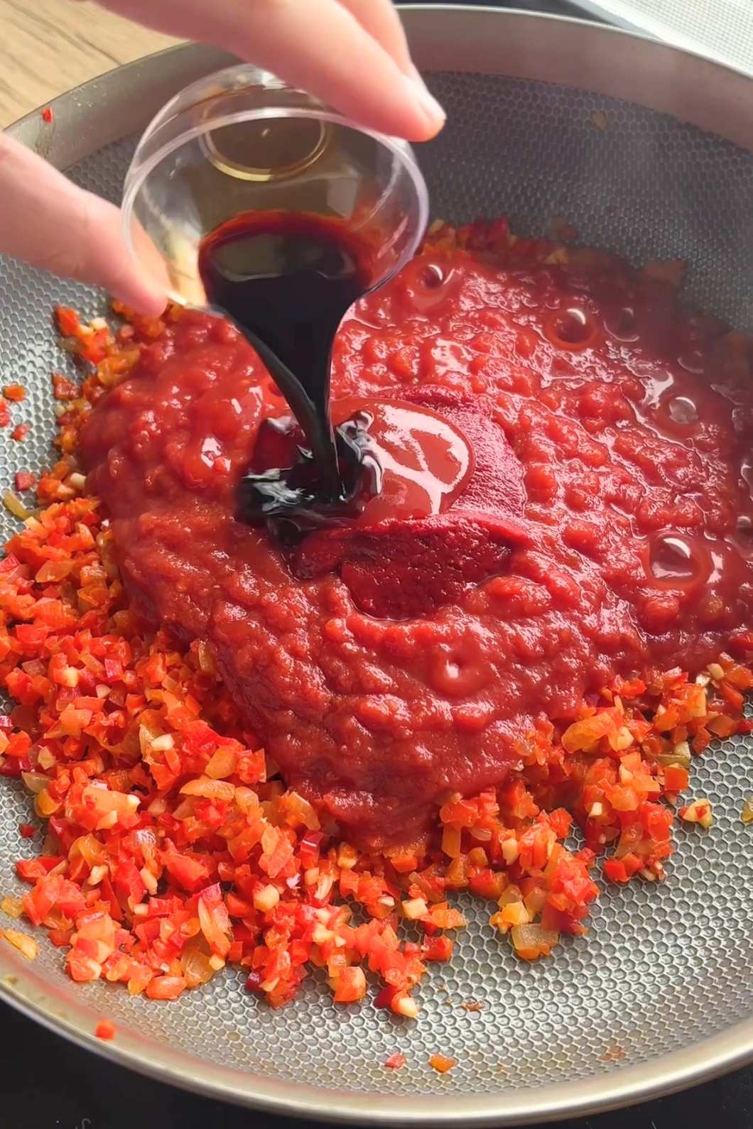 Hand pouring dark sauce over tomato sauce and finely diced red capsicum in a pan.