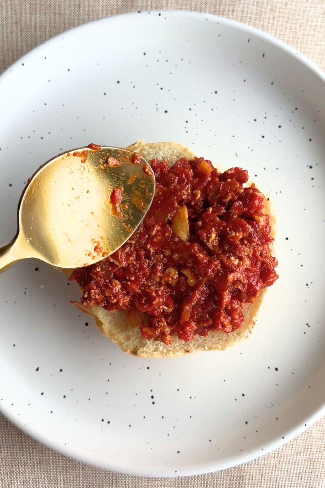 Golden spoon spreading red sauce onto a bun half on a white speckled plate.