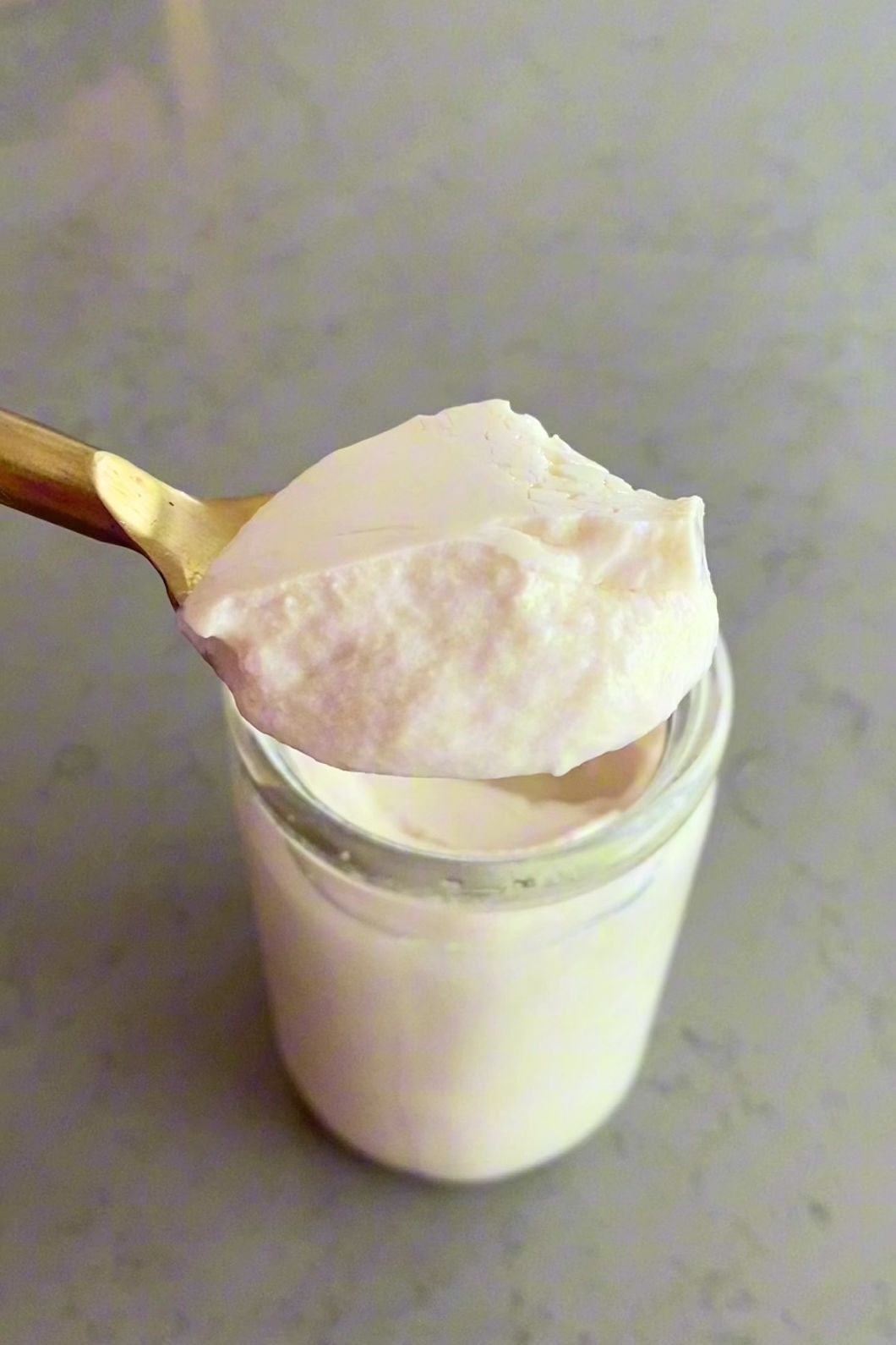 Spoon holding a dollop of thick creamy yogurt above a small glass jar.