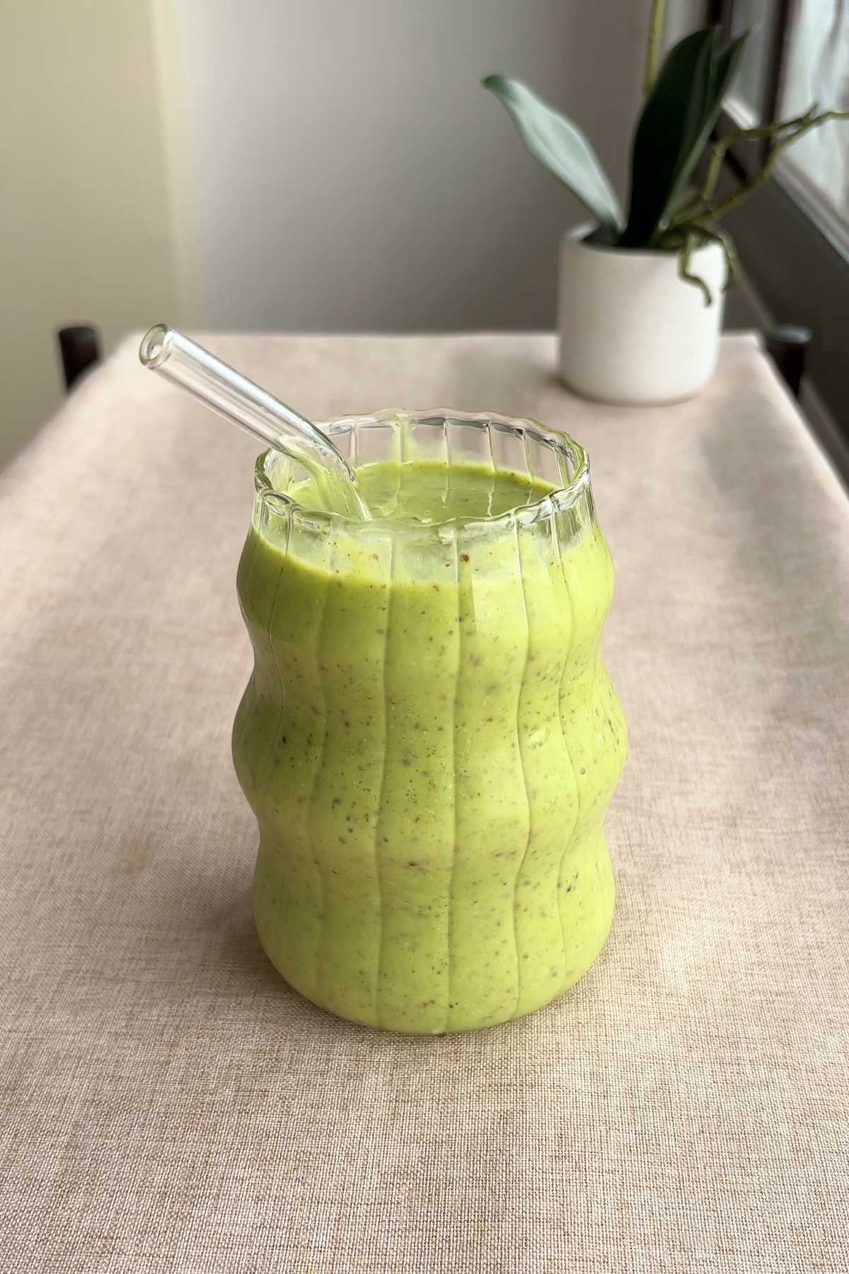 A wavy glass filled with a bright green smoothie and a clear reusable straw resting inside.