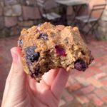 A partially eaten oat cookie studded with blueberries held between fingers over a brick patio.