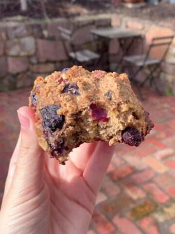 A partially eaten oat cookie studded with blueberries held between fingers over a brick patio.