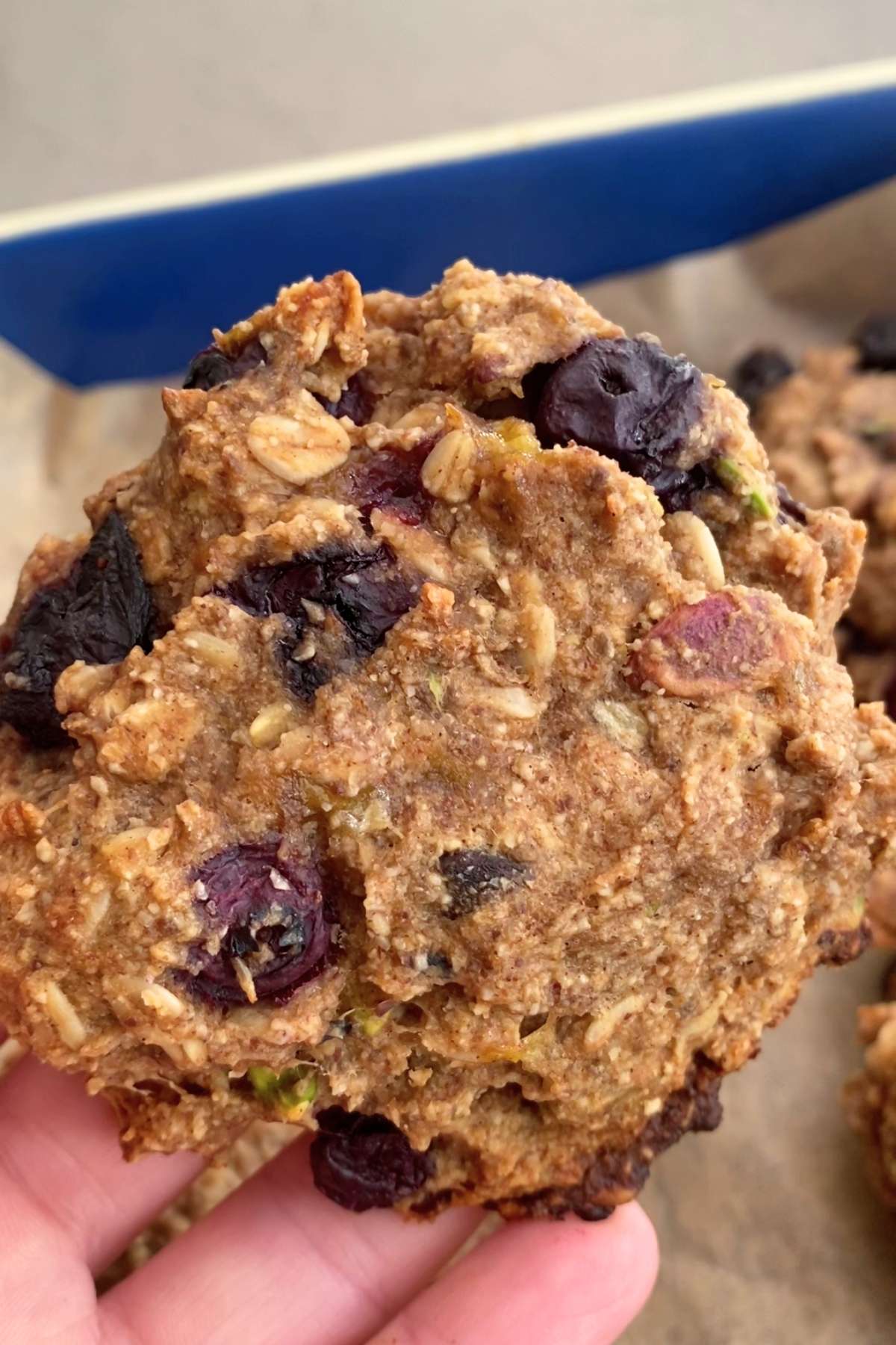A hand holding a rustic, rounded oat cookie with visible blueberry pockets.