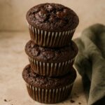 Three chocolate muffins stacked on a neutral surface with a green cloth beside them.