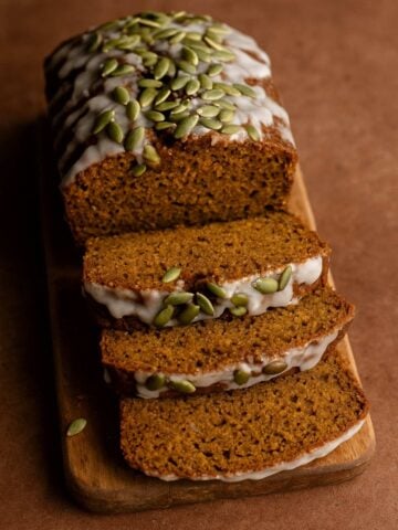 Sliced pumpkin loaf with white glaze and pepitas on top, arranged on a wooden board.