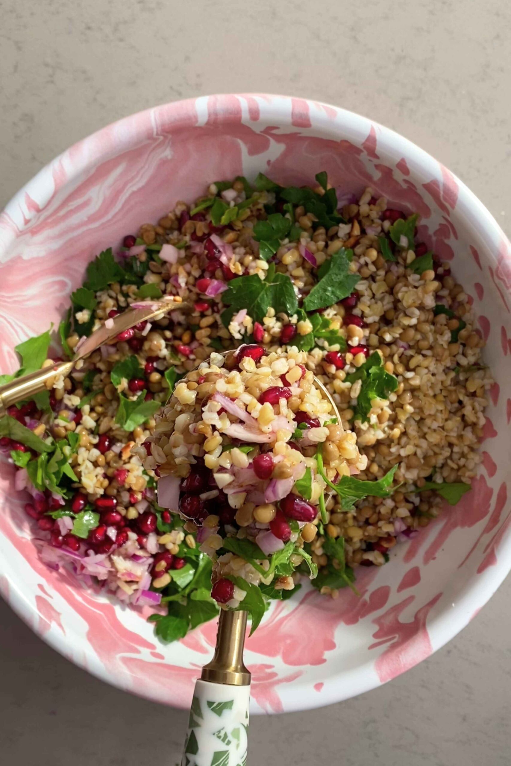 A gold spoon lifting grains, parsley leaves, and pomegranate seeds from a pink-and-white marbled bowl.