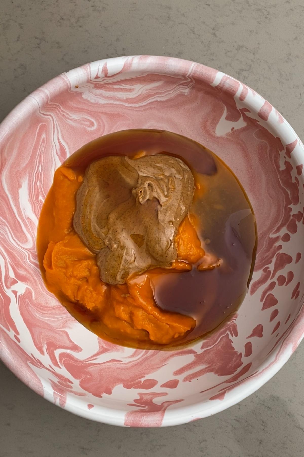 Almond butter, maple syrup, and pumpkin purée sitting unblended in a pink marbled mixing bowl.