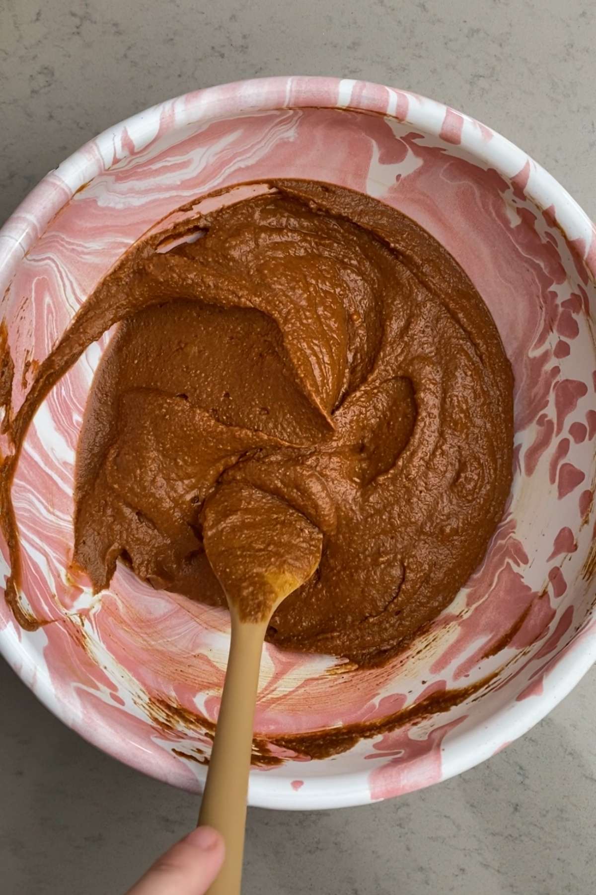 Thick pumpkin brownies batter stirred with a beige spatula in a pink marbled mixing bowl.