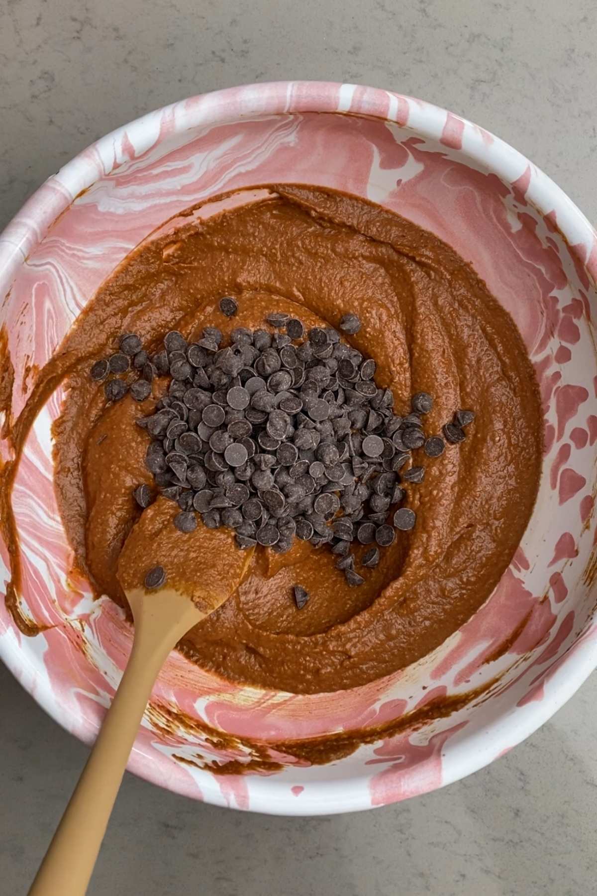 Chocolate chips piled on top of thick pumpkin brownie batter in a pink marbled mixing bowl with a beige spatula.