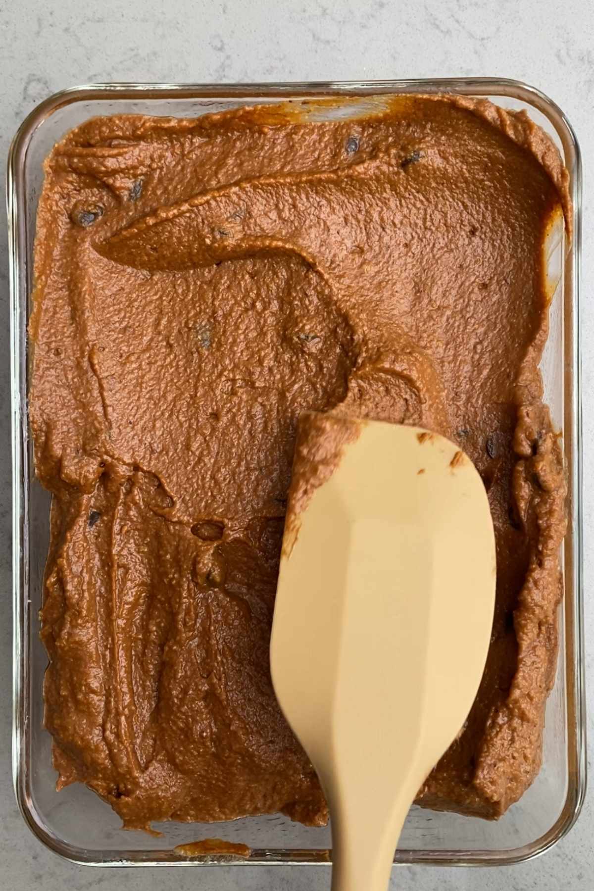 Thick pumpkin brownie batter being spread evenly in a glass baking dish using a beige spatula.