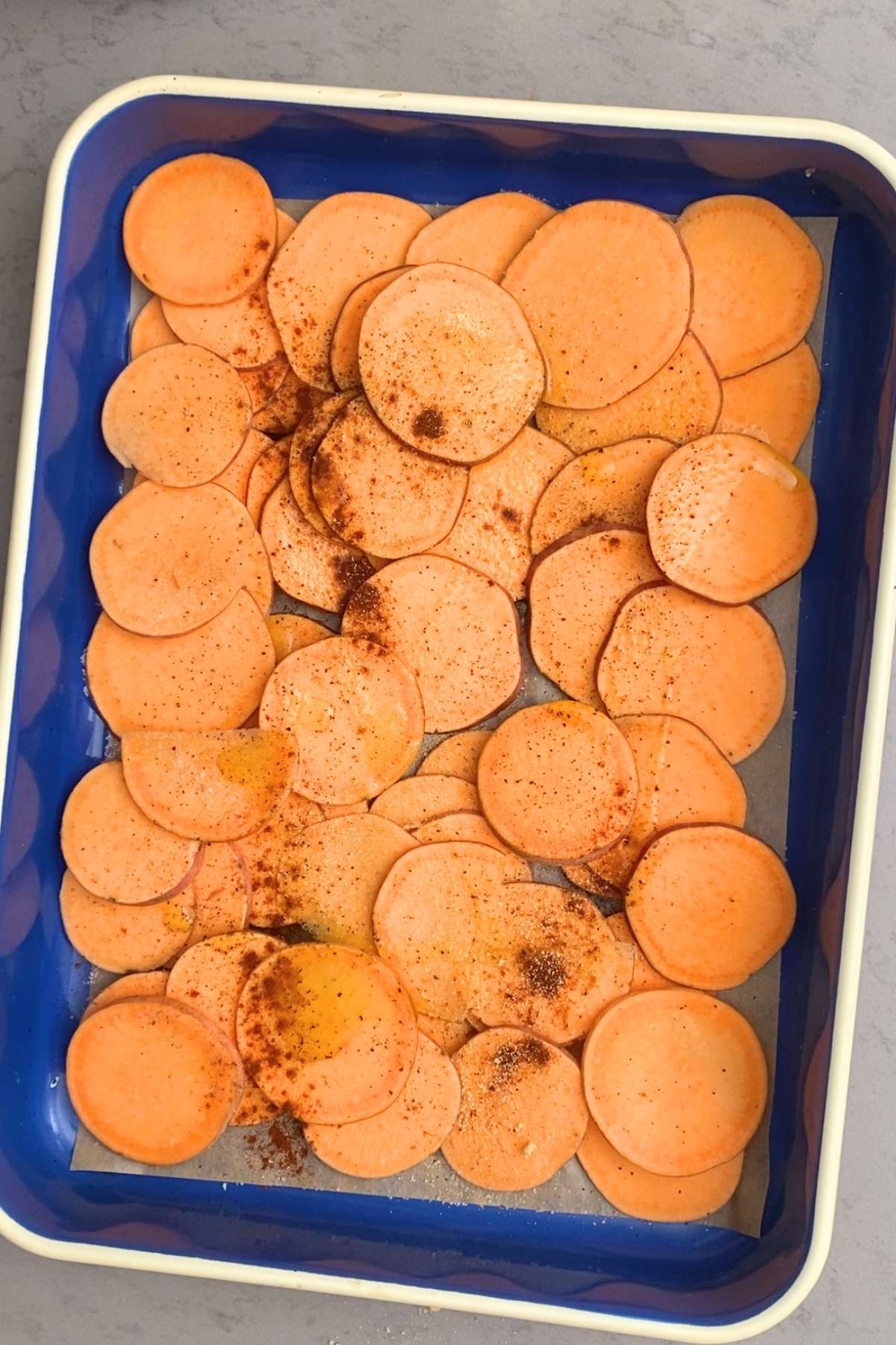 Thinly sliced sweet potatoes sprinkled with spices in a blue roasting dish.