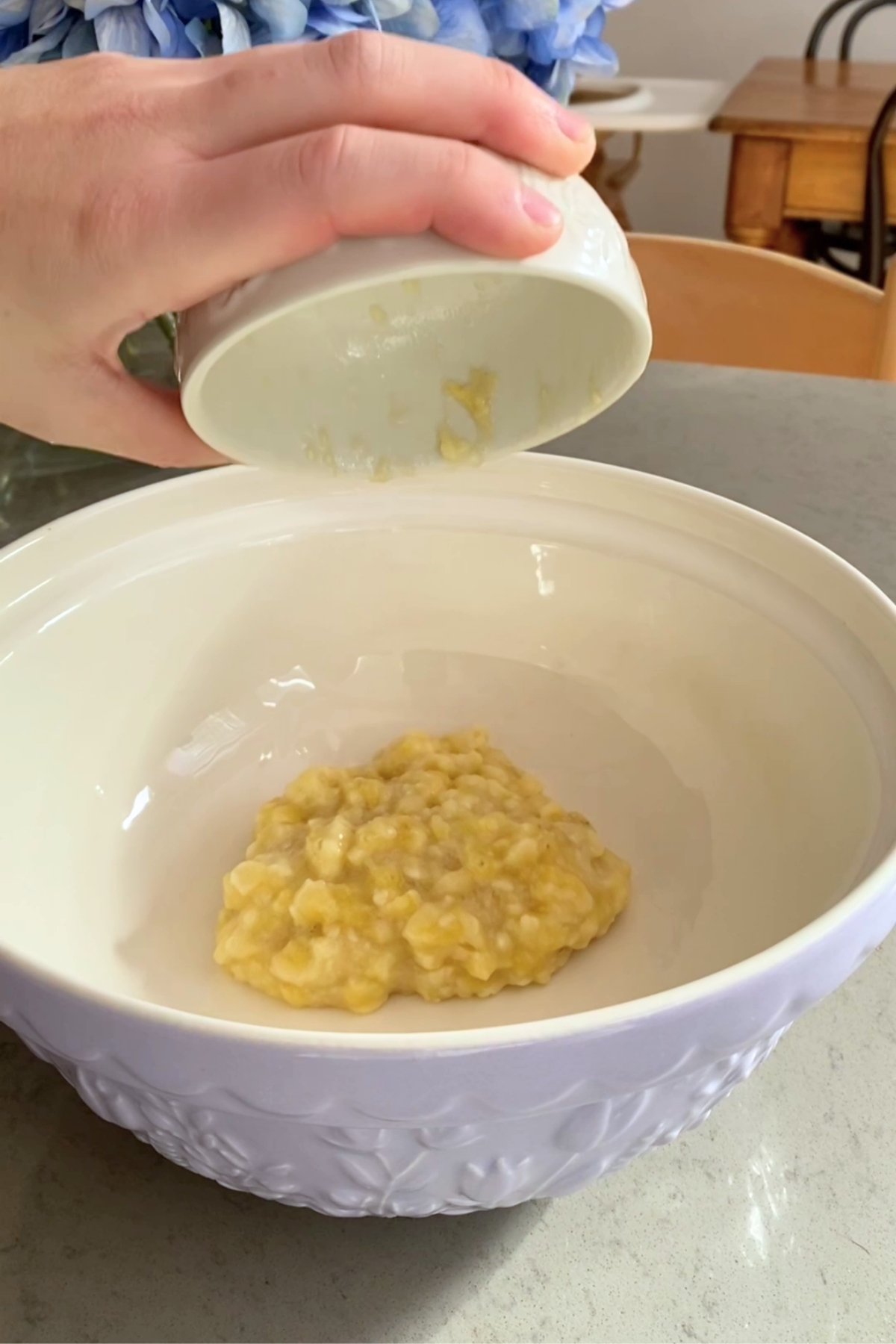 A hand tips a small bowl to dump mashed bananas into a large white mixing bowl.