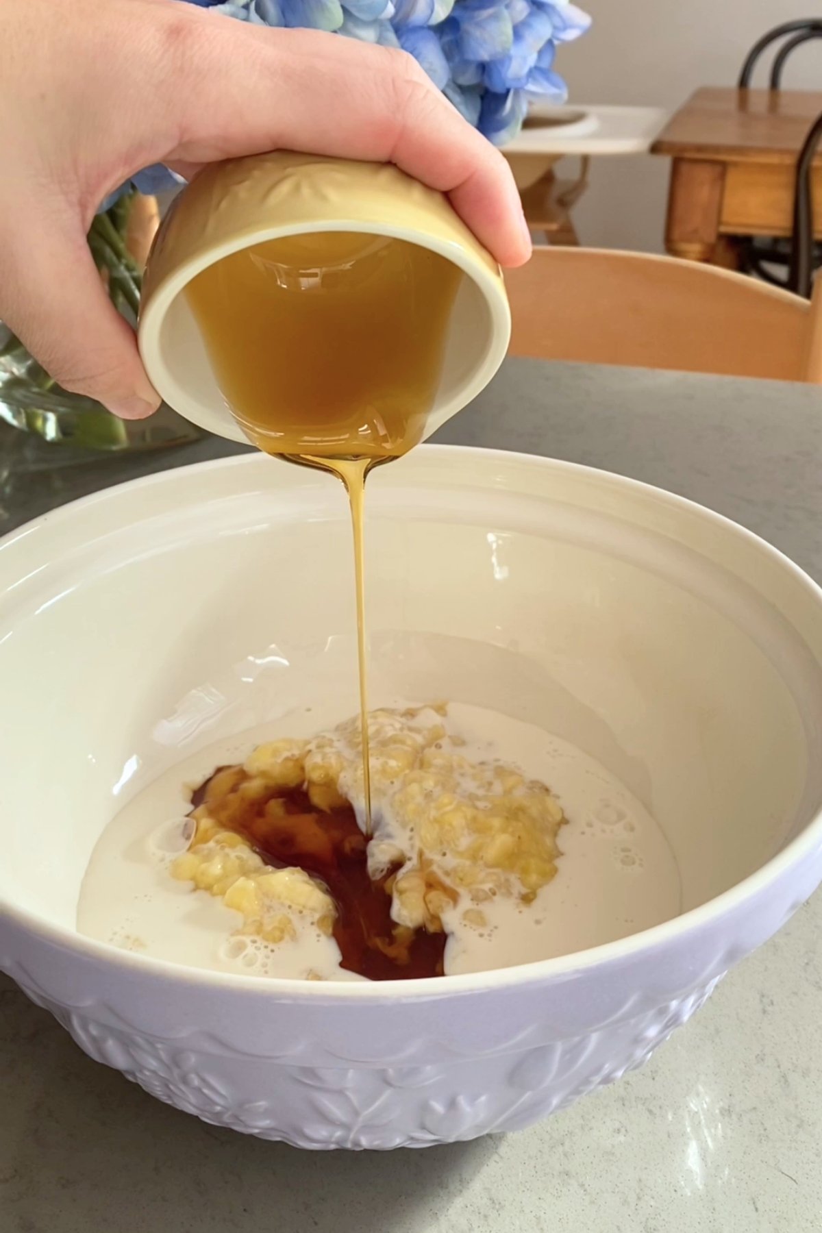 A hand tips a small bowl to dump mashed bananas into a large white mixing bowl.