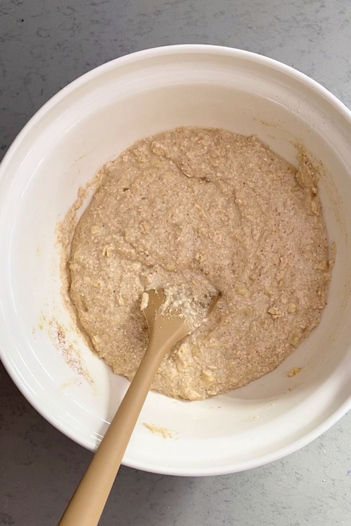A spatula sits in a white bowl of thick, speckled oat batter.