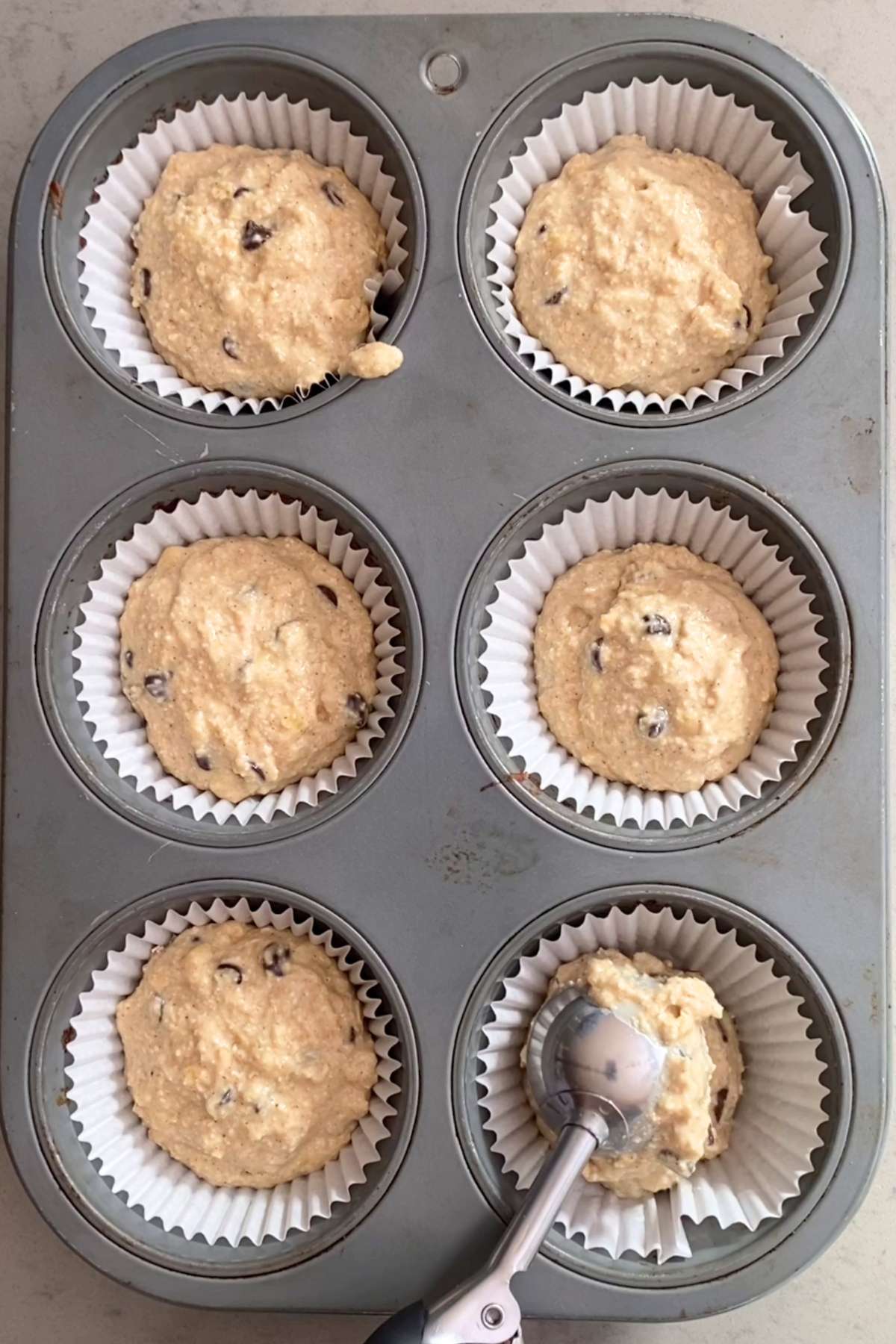 Six unbaked batter mounds fill paper liners in a metal muffin tin, with a scoop adding more.