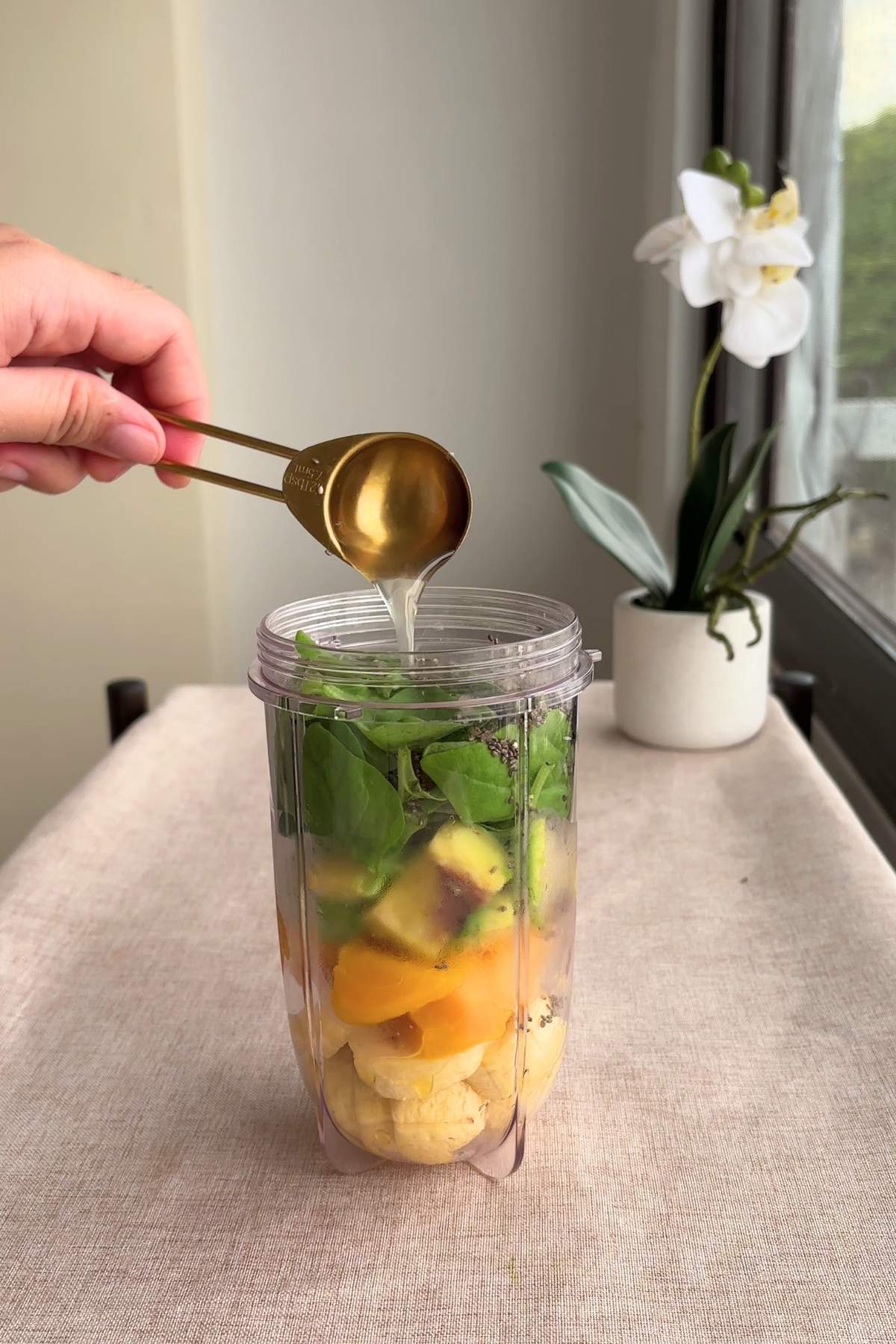 A gold measuring spoon drizzling liquid into the blender cup filled with spinach and fruit.