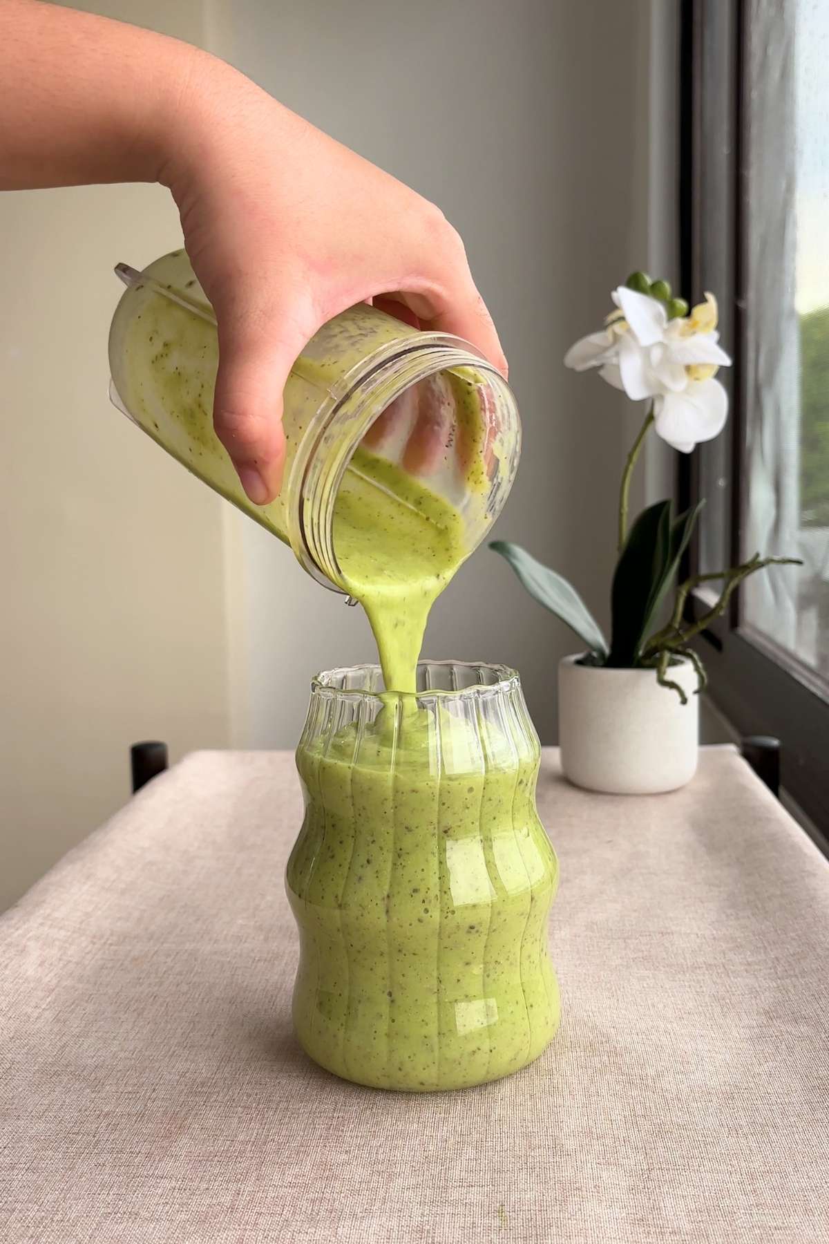A hand tilting the blender cup to pour the finished green smoothie back into the wavy glass.