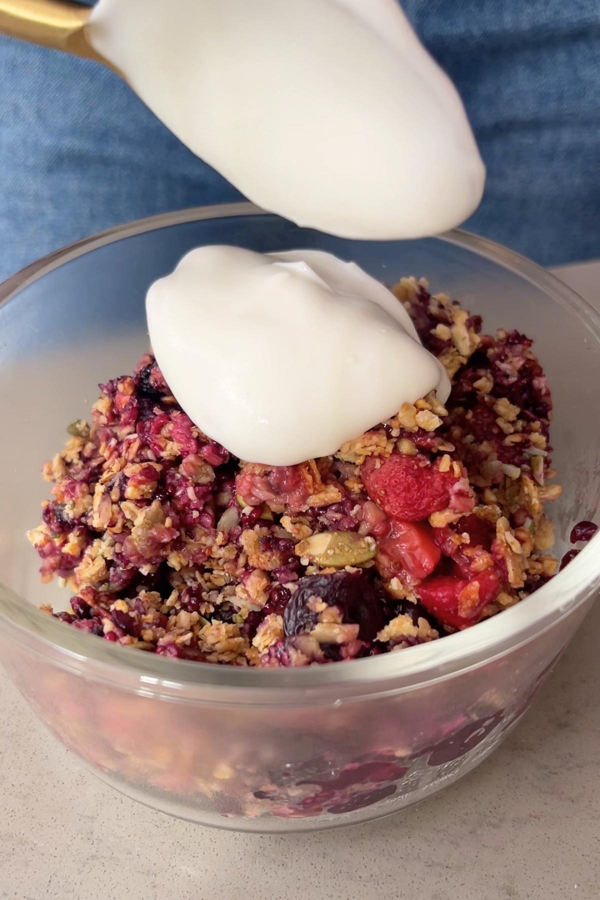 Spoon dolloping yogurt onto a serving of baked berry crumble in a bowl.