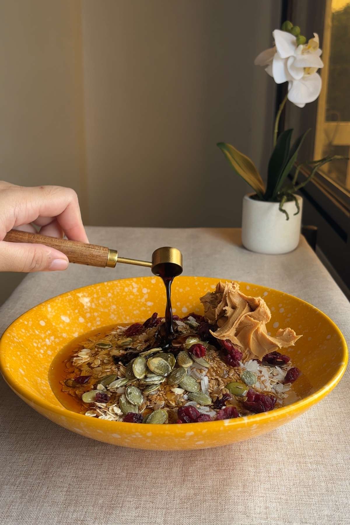 A spoon drizzling thick syrup over oats, cranberries, pepitas, and a scoop of nut butter in a yellow bowl.