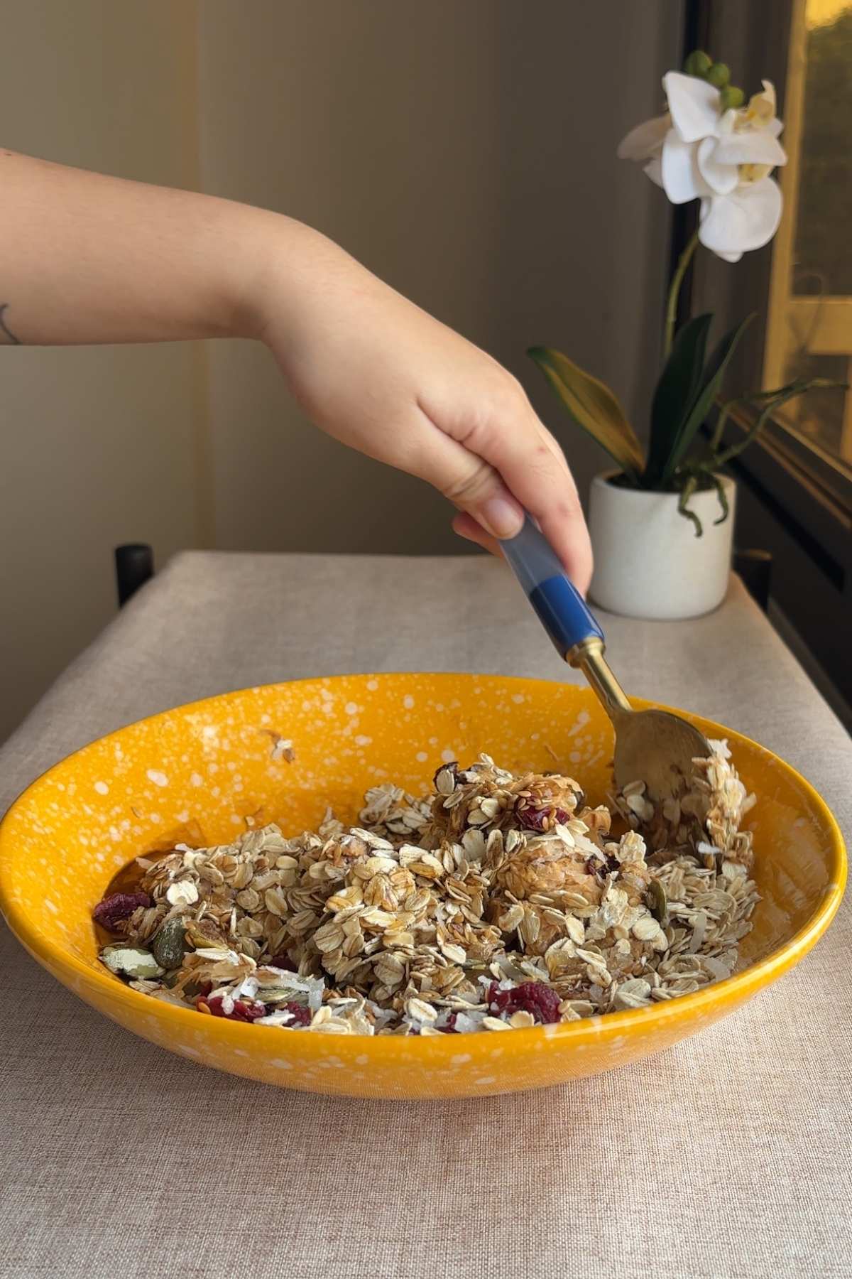Hand mixing oat mixture with a spoon inside a large yellow bowl on a beige surface.