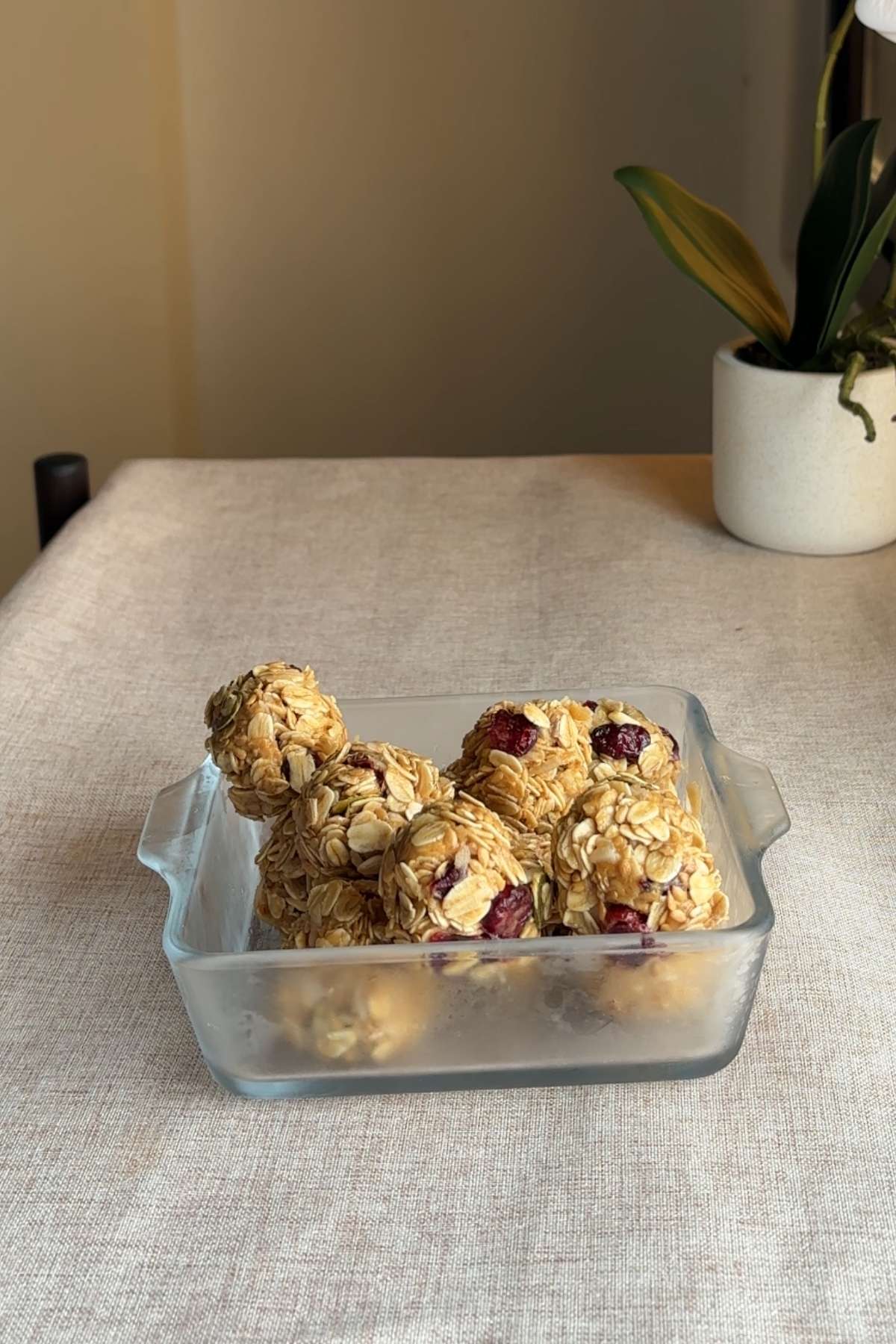 Glass container filled with oat and cranberry balls stacked on a beige cloth surface.