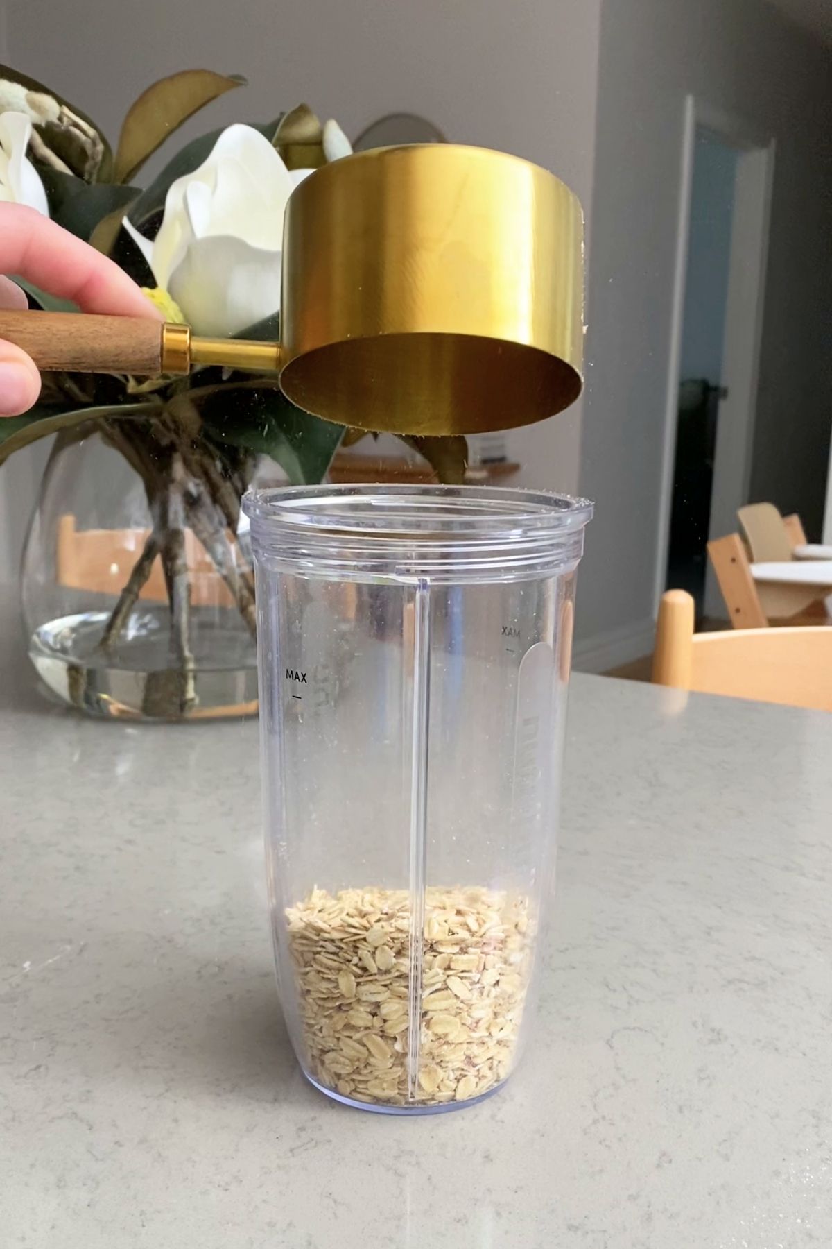 A brass measuring cup pouring rolled oats into a clear blender cup marked with a max fill line.