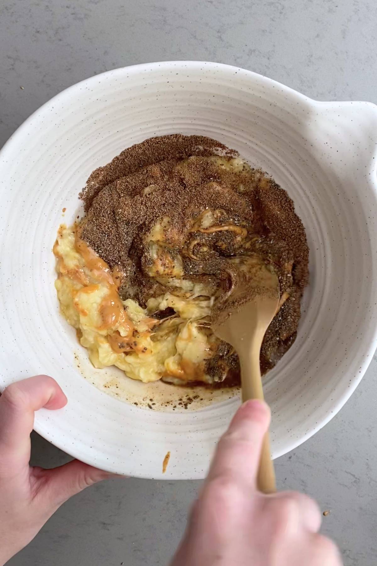 A hand stirring mashed banana, flaxseed, and chia in a white bowl with a gold spoon.