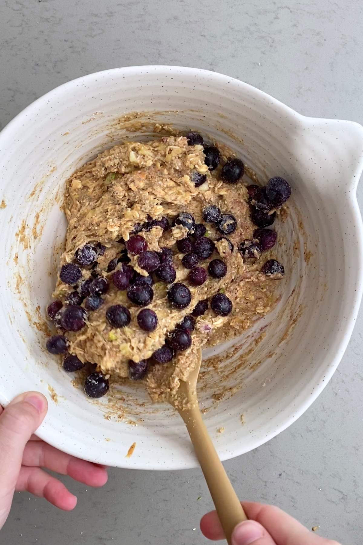 A spoon folding frozen blueberries into a coarse oat-and-nut mixture in a mixing bowl.