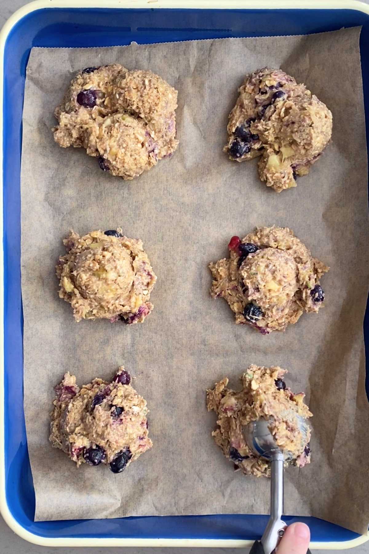 A cookie scoop dropping oat-blueberry dough onto parchment paper in a rectangular pan.
