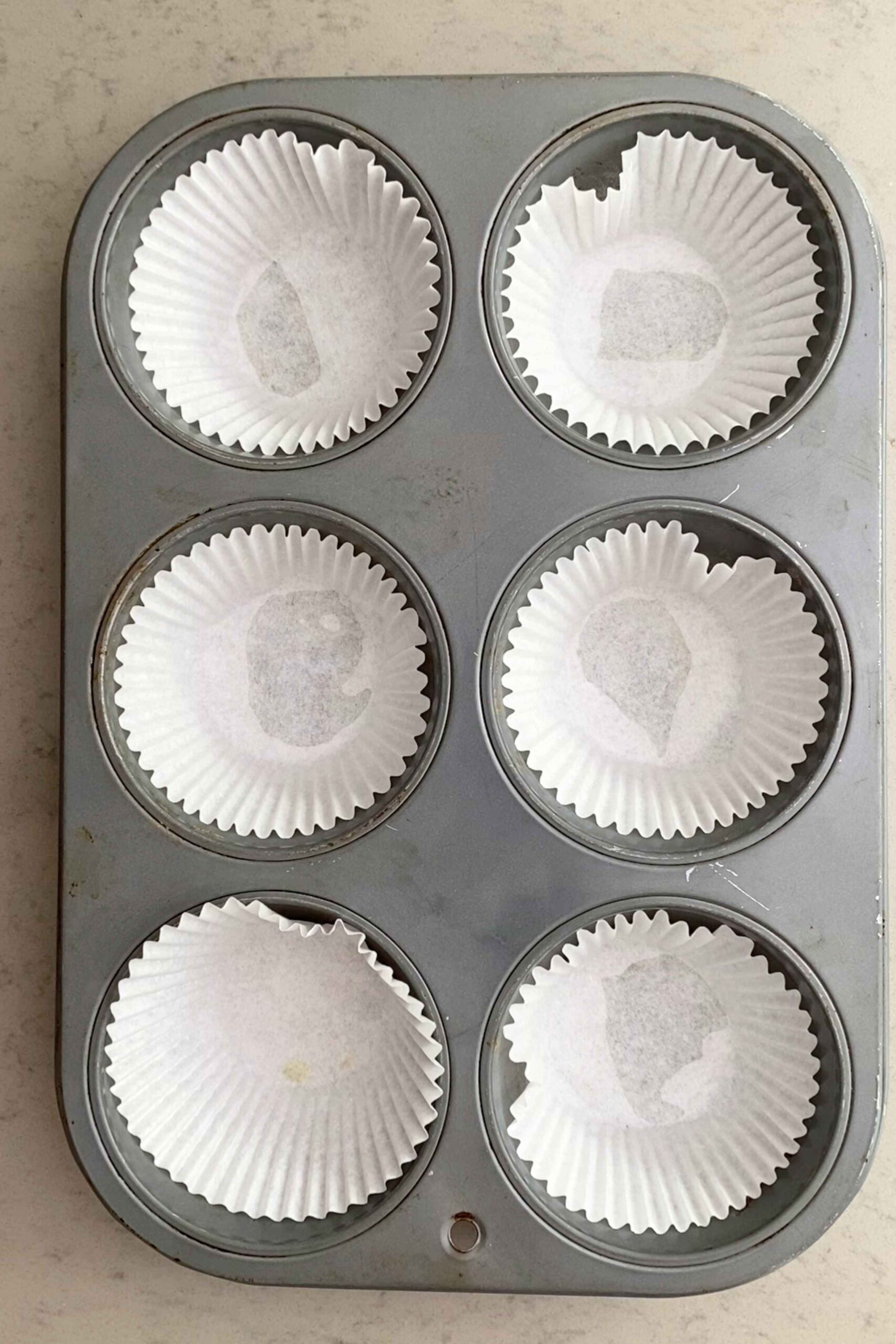 Muffin tin with six white paper liners neatly placed in each cup.