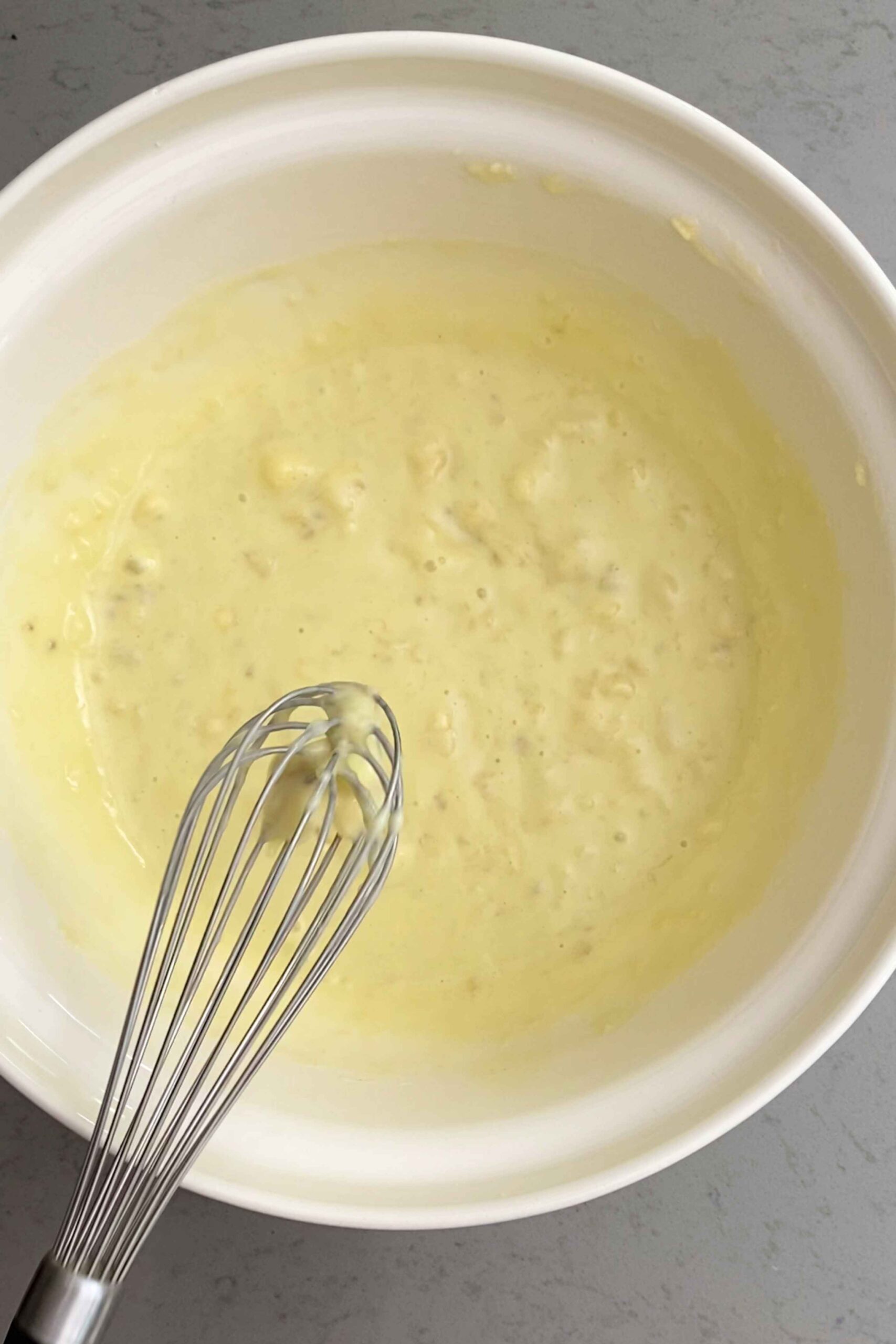 Whisk resting in a bowl of pale, lumpy banana-based wet batter.