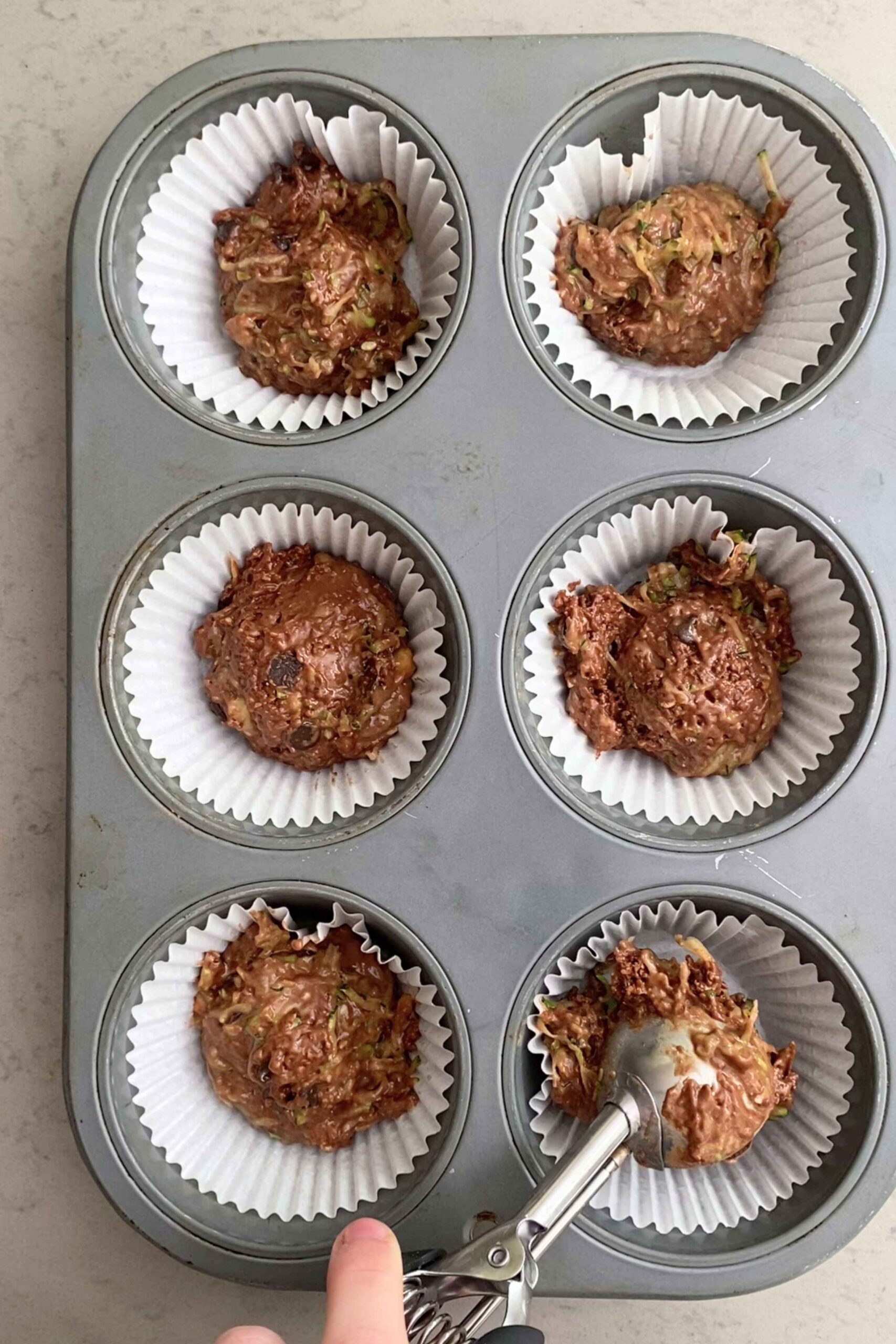 Ice-cream scoop depositing zucchini–chocolate batter into a lined muffin cup.
