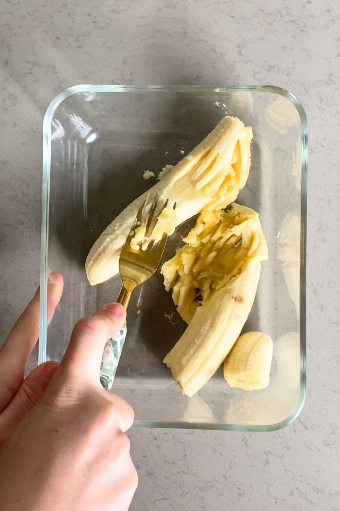 A hand mashing ripe bananas with a fork in a clear glass dish on a light gray counter.