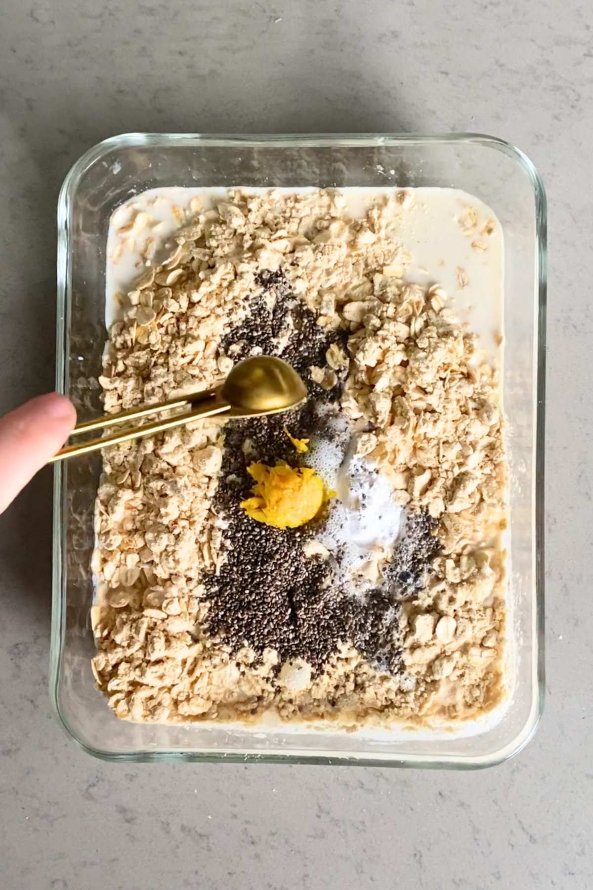 Dry and wet ingredients, including oats and chia seeds, being added to a glass dish with a measuring spoon.