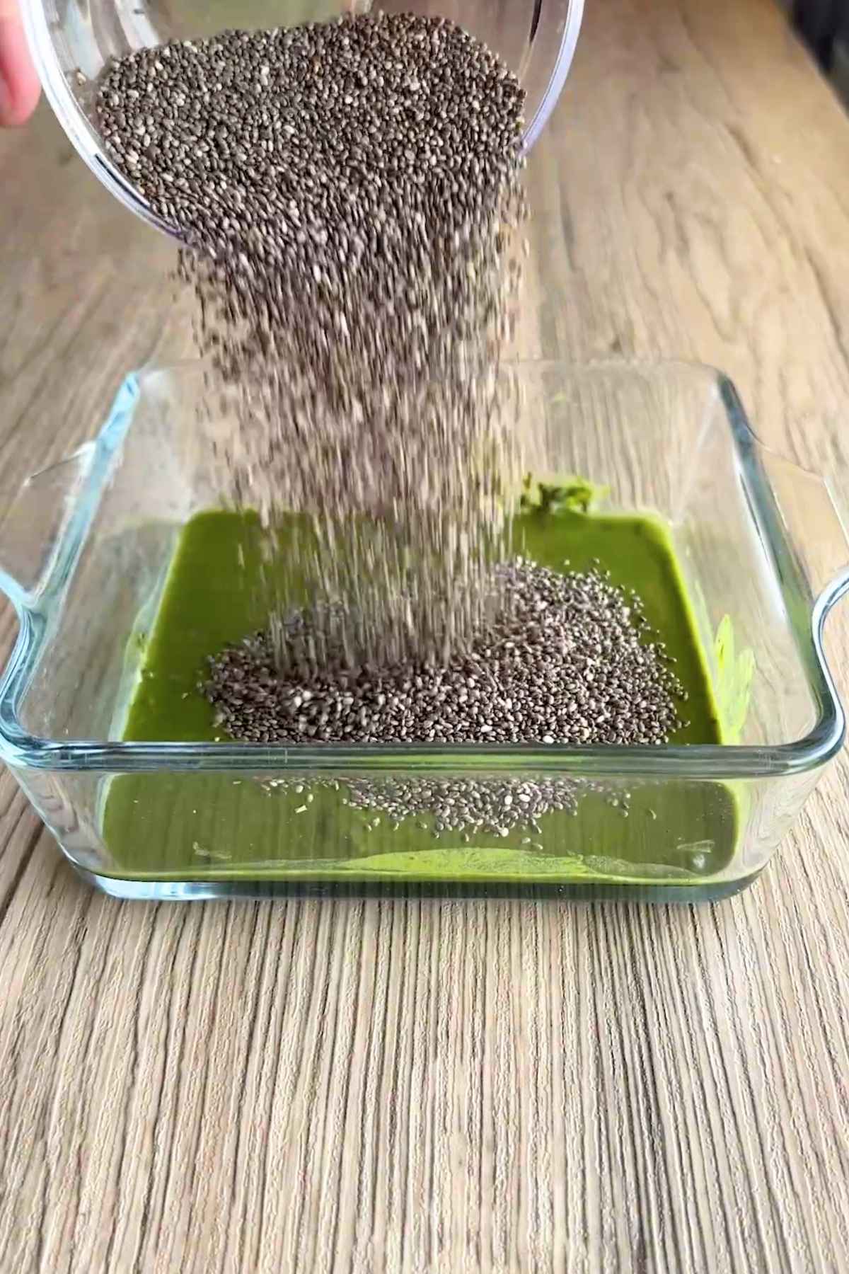 Chia seeds pouring onto green matcha mixture in a glass dish.