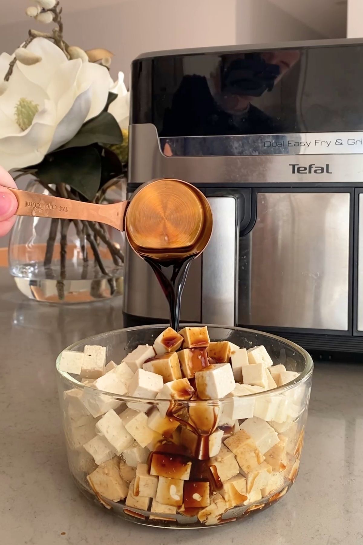 A copper measuring cup pours dark sauce over a bowl of small tofu cubes.