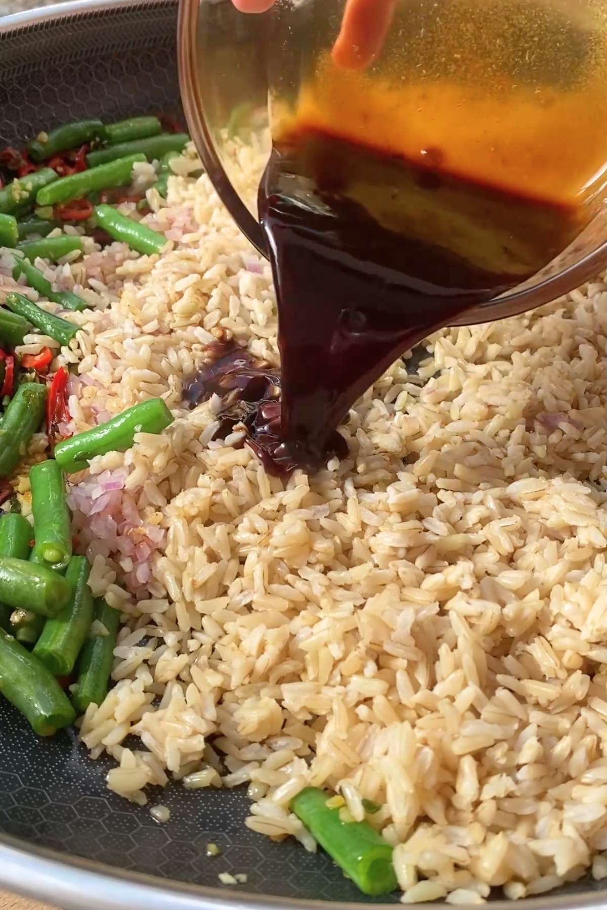 Light brown rice in the pan being coated with dark sauce beside green beans.