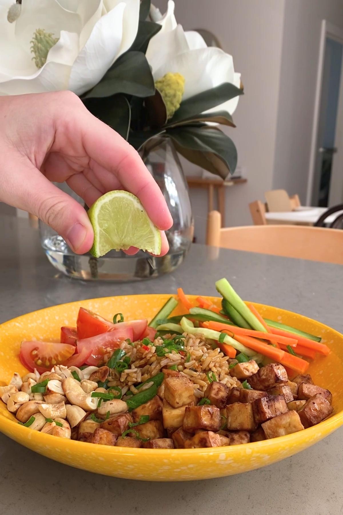 A hand squeezes a lime wedge over a bowl of rice topped with crispy tofu cubes, carrot and cucumber ribbons, tomato wedges, and cashews.