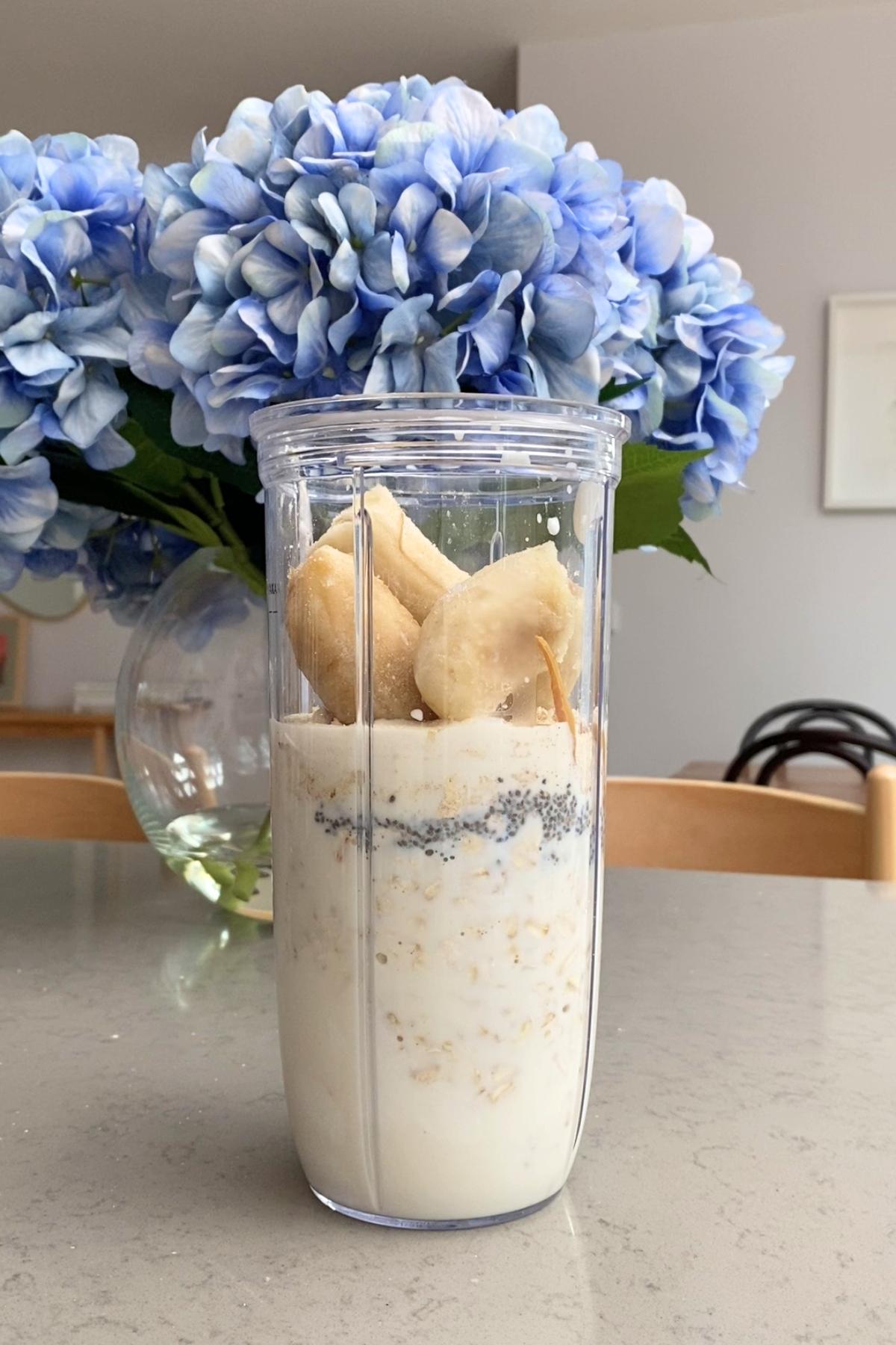 Clear blender cup with plant milk, oats, chia seeds and frozen banana chunks.