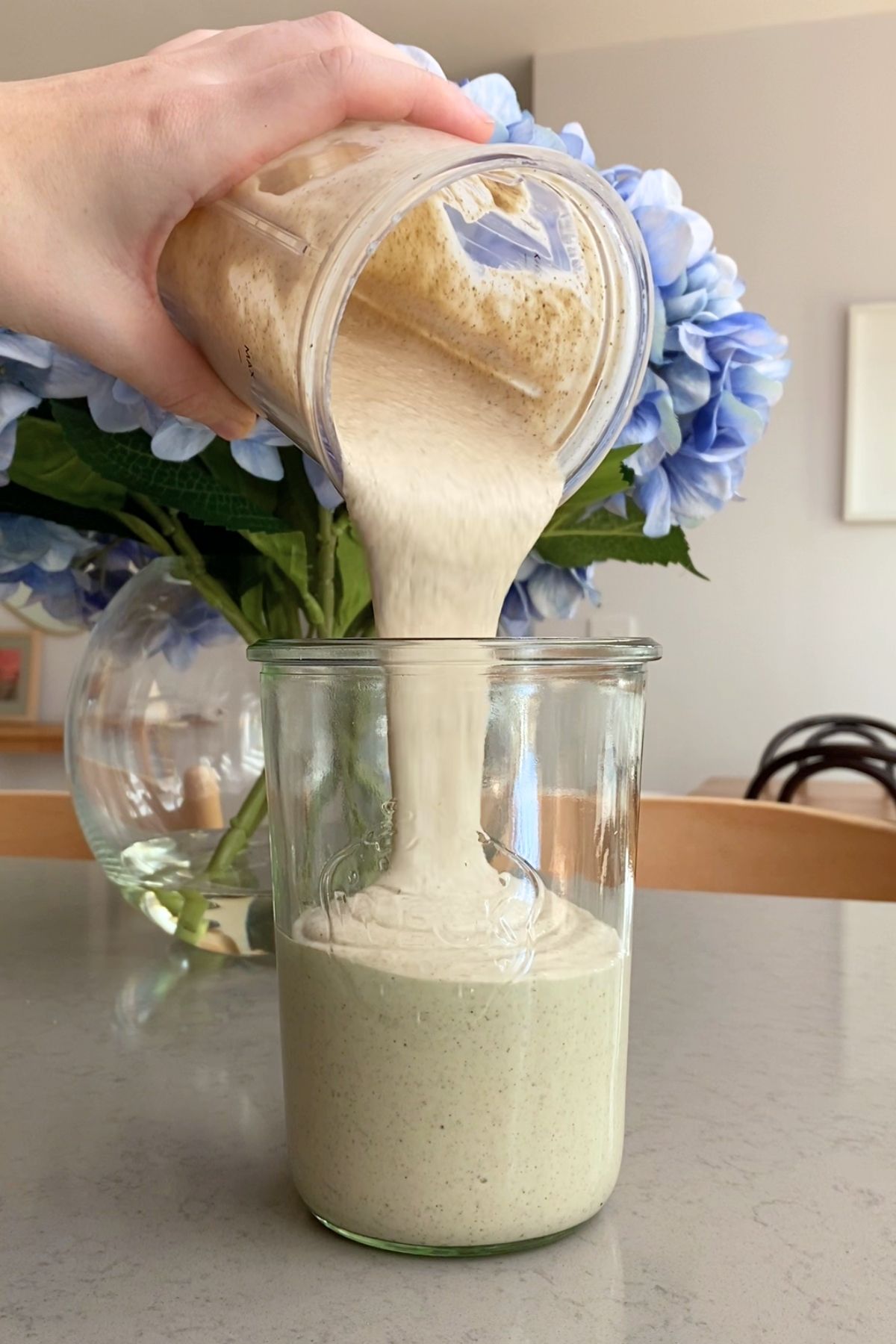 Smooth beige smoothie pouring from blender container into a tall glass jar.