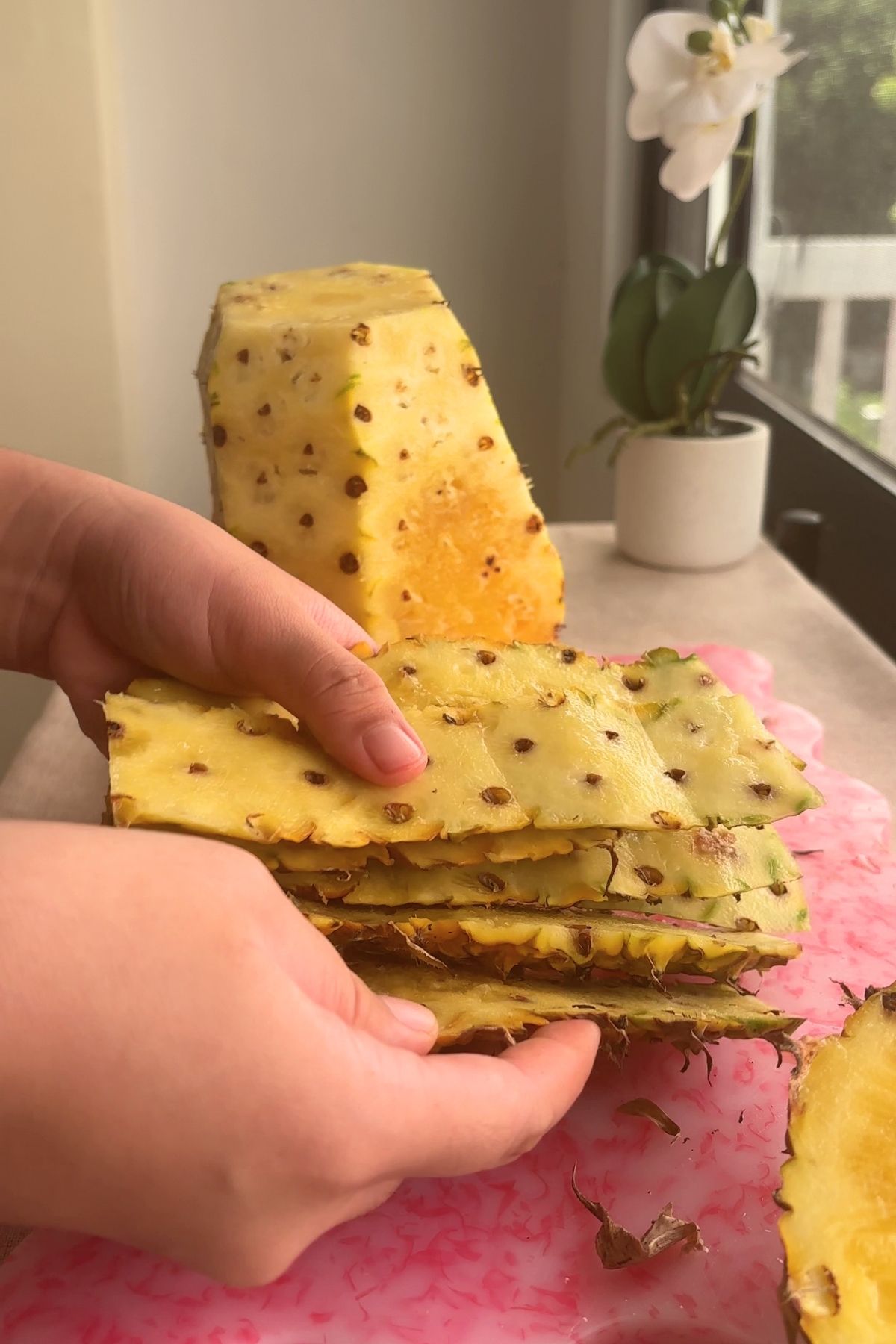 Hands holding peeled pineapple skins beside a cored pineapple on a pink board.