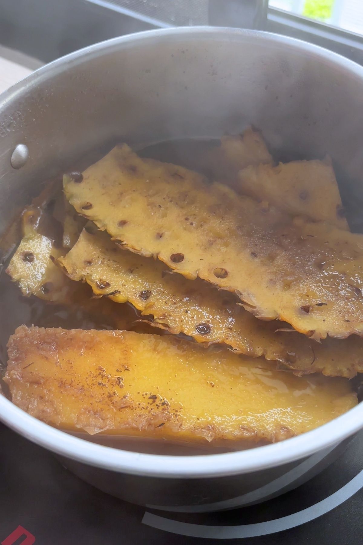 Flattened pineapple skins floating in dark simmering liquid inside a saucepan.