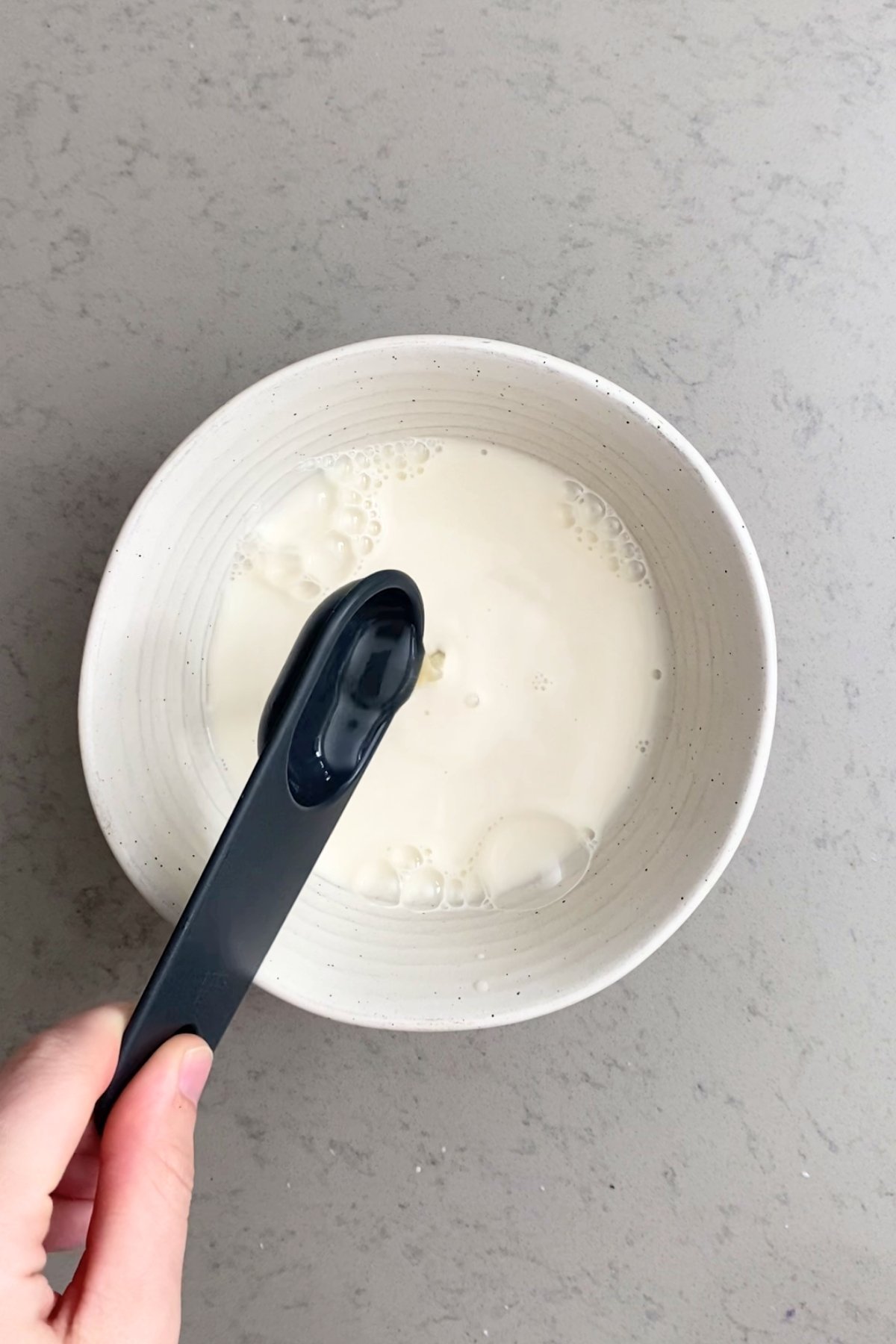 Vinegar being added to soy milk in a white bowl using a black measuring spoon.