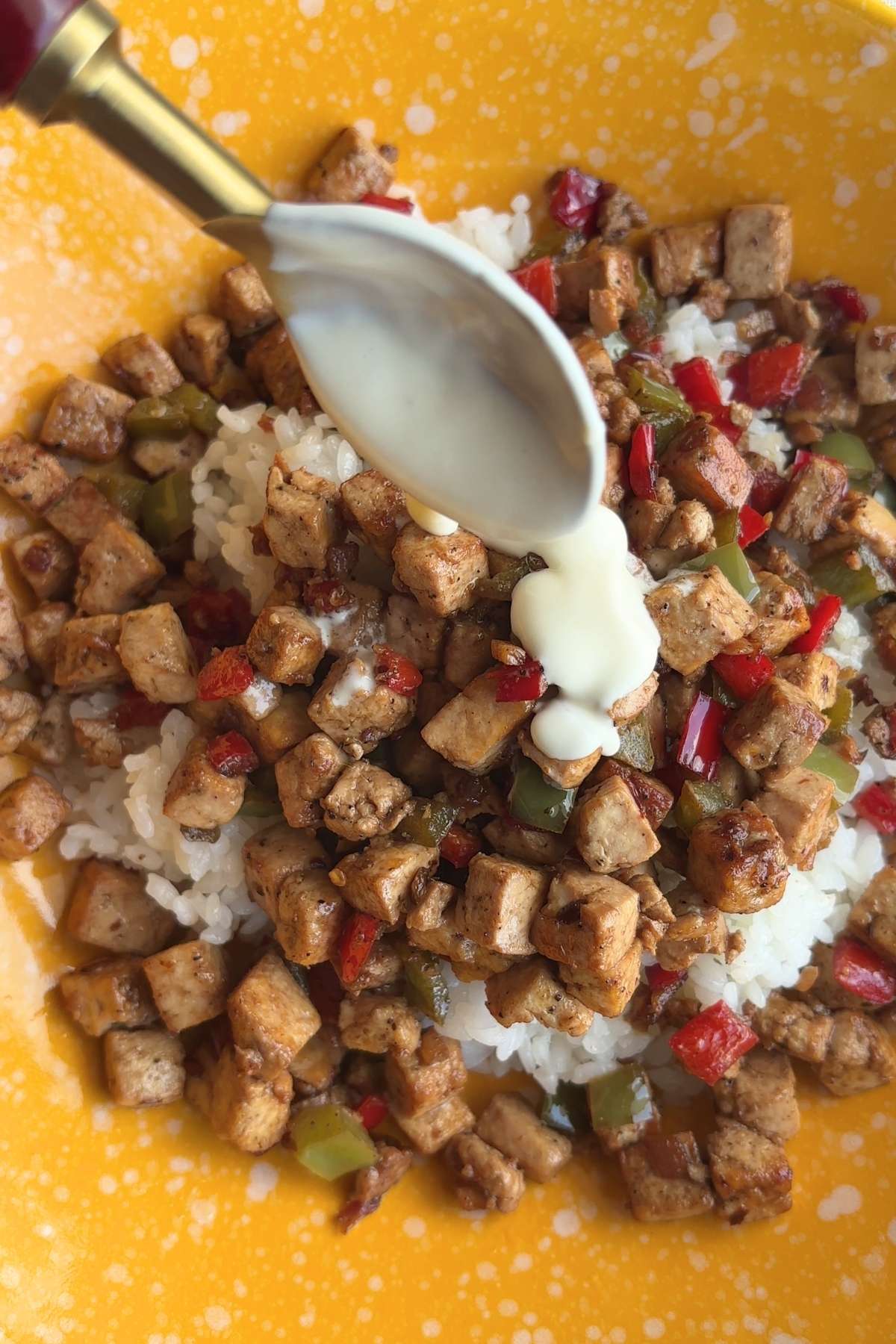 Creamy sauce drizzled over crispy tofu cubes and diced red and green peppers atop a bed of white rice in a yellow bowl.